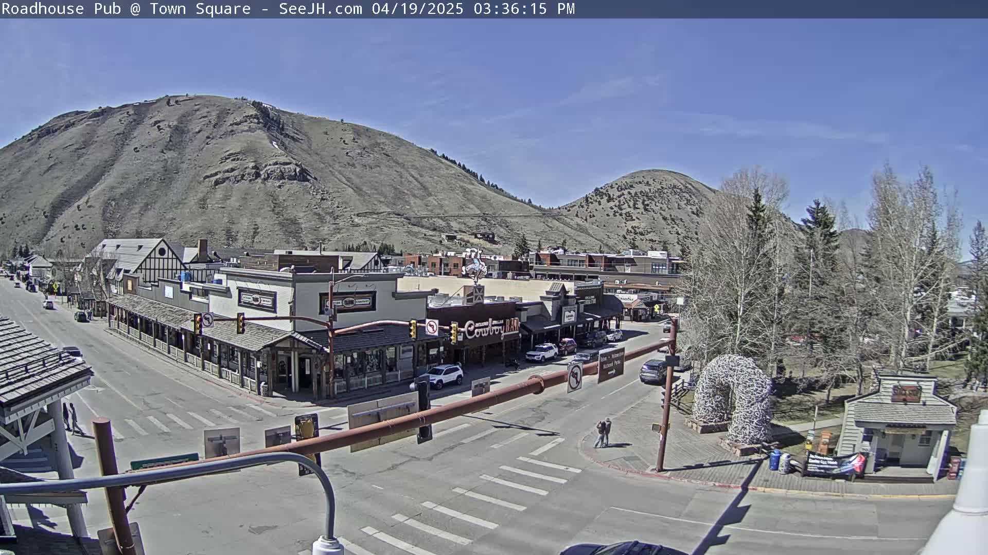 A sunny, daytime view of a town square nestled between two mountains, featuring several buildings, a street with cars, and a park-like area with trees.