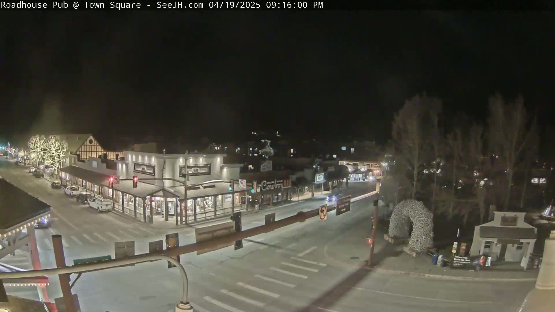 A nighttime view of a town square with several buildings and sparsely trafficked streets, lightly snow-covered.