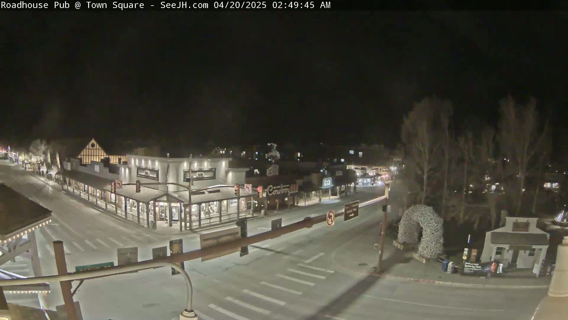 A snow-covered town square at night, illuminated by streetlights, shows several buildings and a sparse amount of traffic.