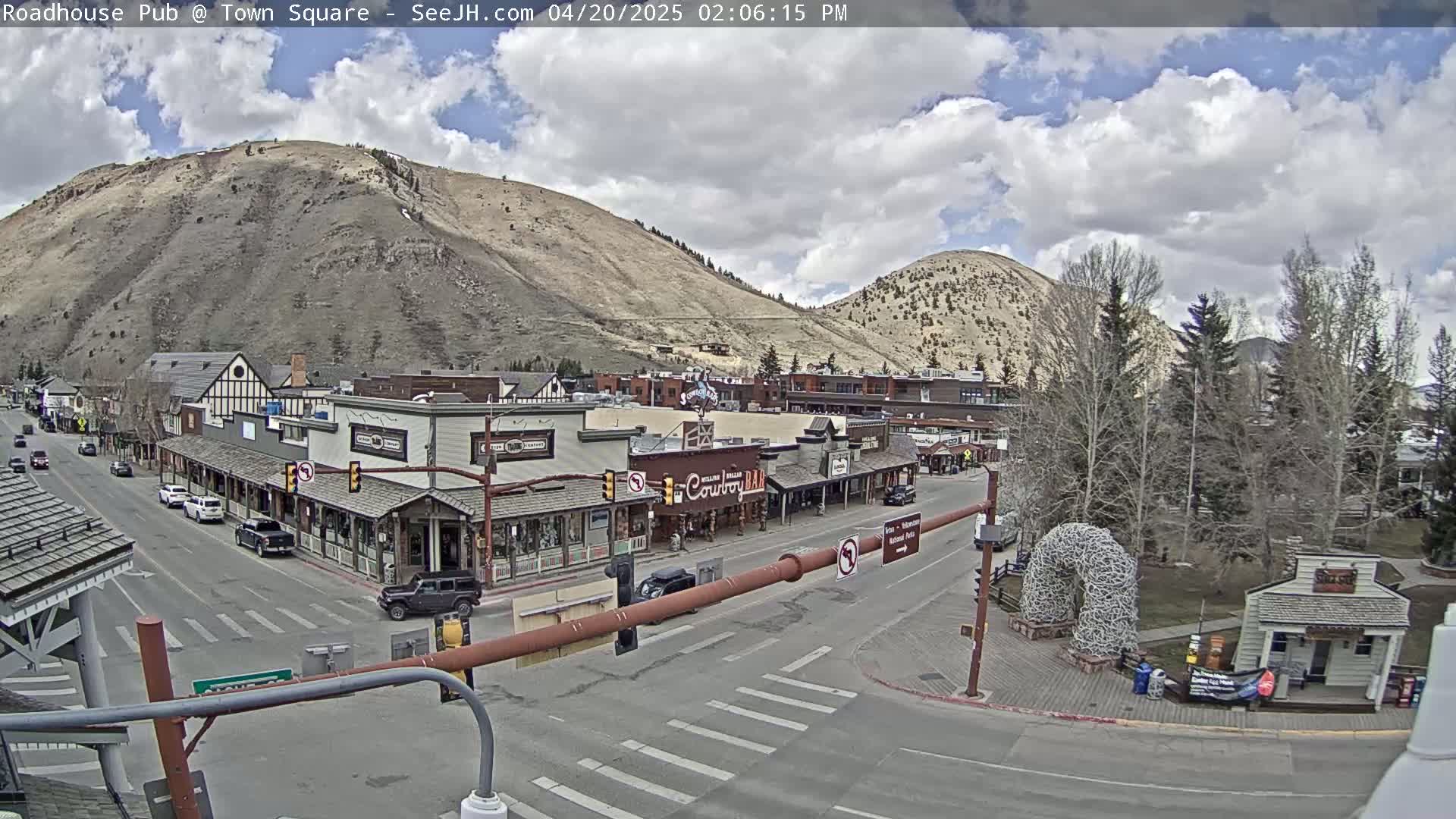 A partly cloudy day reveals a town square nestled between two hills, with several shops and a few vehicles visible on the streets.