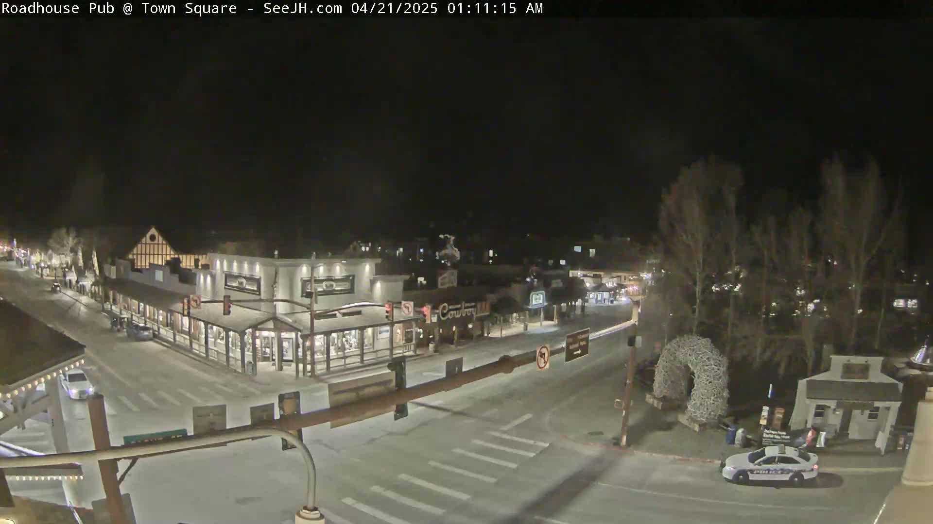 A nighttime, snow-covered town square with several buildings, a police car, and sparsely populated streets.