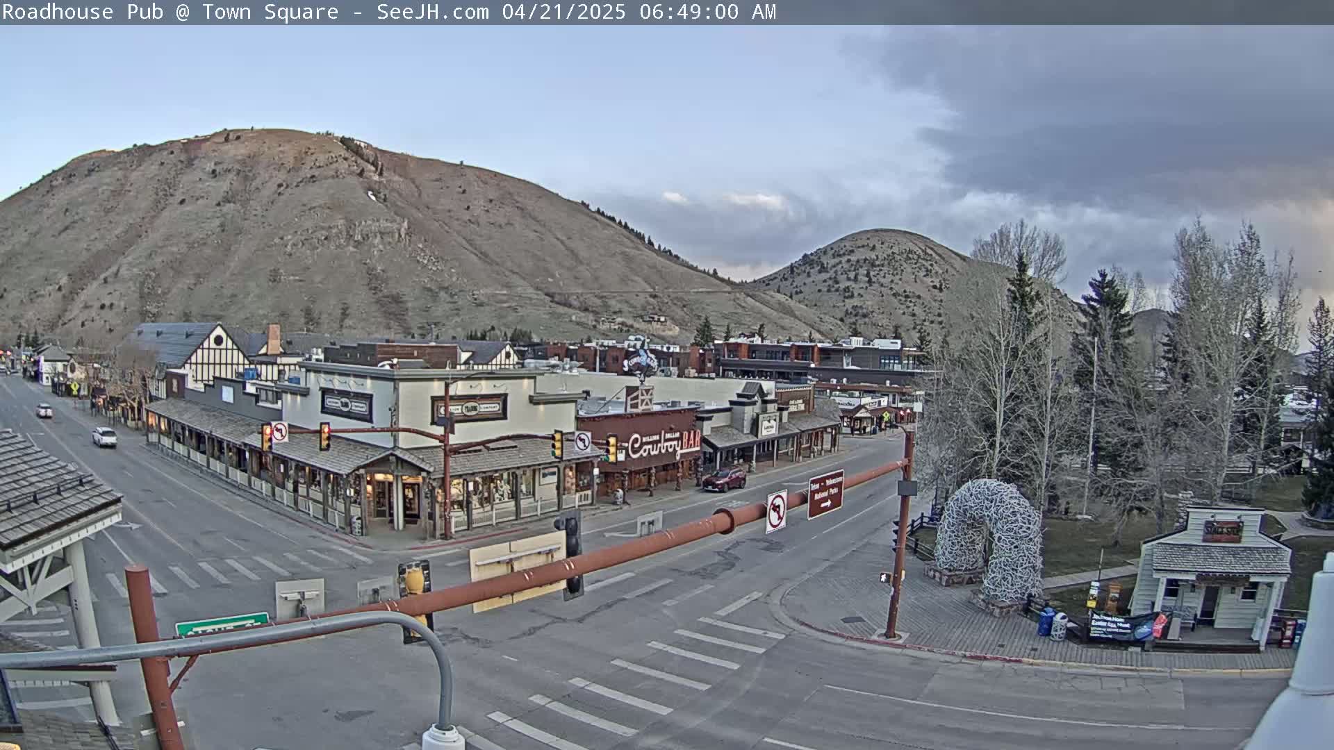 A mostly cloudy morning reveals a mountain-backed town square with several storefronts and sparse vehicle traffic.