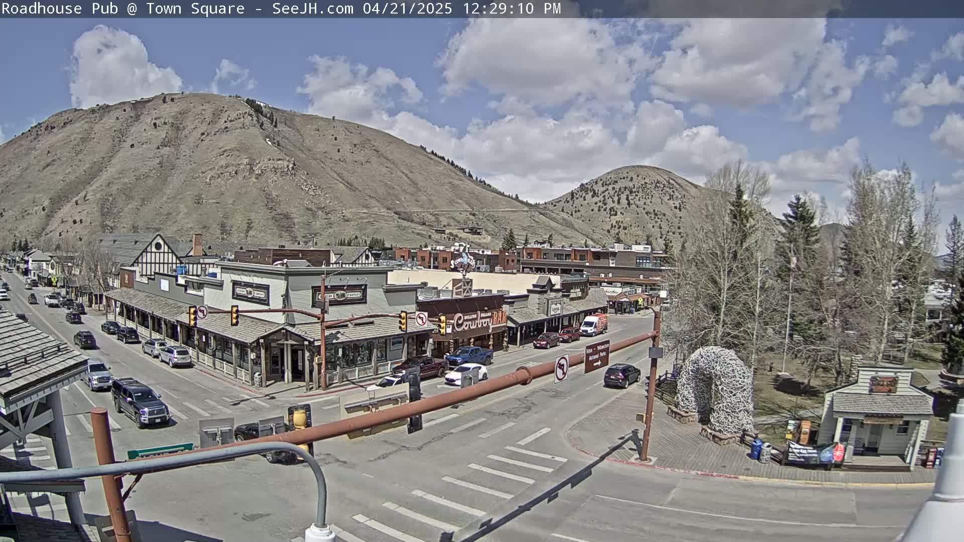 A partly cloudy day shows a town square with cars parked and driving on roads that wind past buildings in a mountainous region.