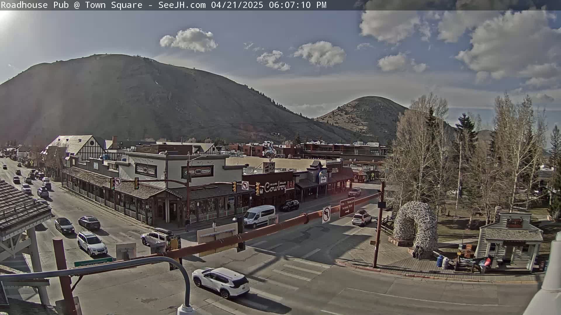 A sunny day reveals a town square scene with buildings, vehicles, and trees, nestled against the backdrop of mountains under a partly cloudy sky.