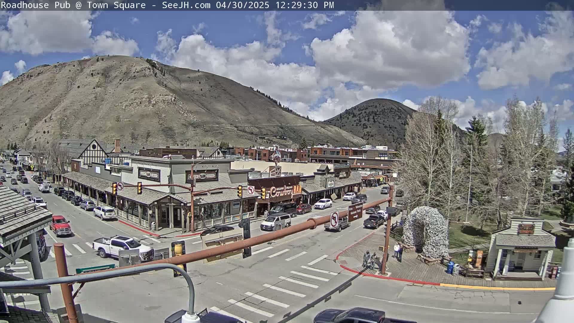 A partly cloudy day reveals a mountainside town square with cars parked along the streets and buildings lining both sides, under a mostly sunny sky.