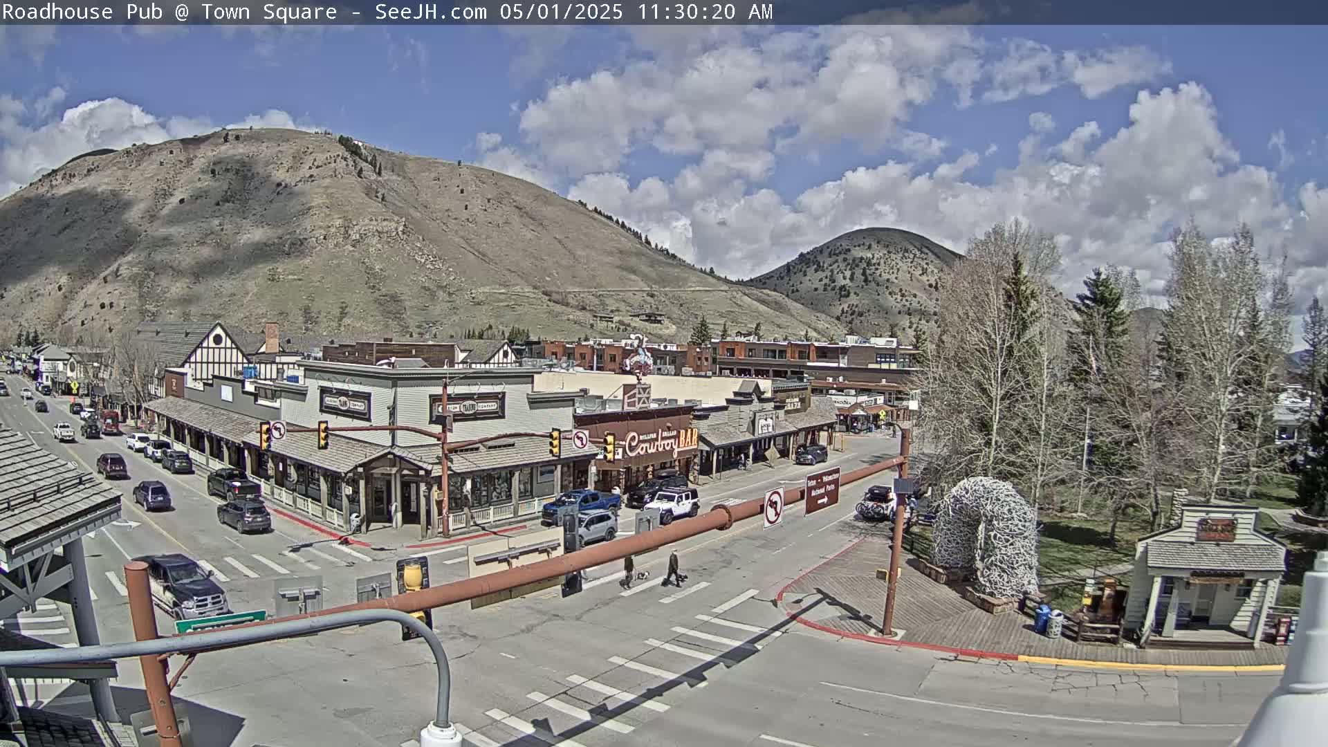 A partly cloudy day reveals a town square nestled between a mountain and trees, with cars parked along the streets and pedestrians crossing.