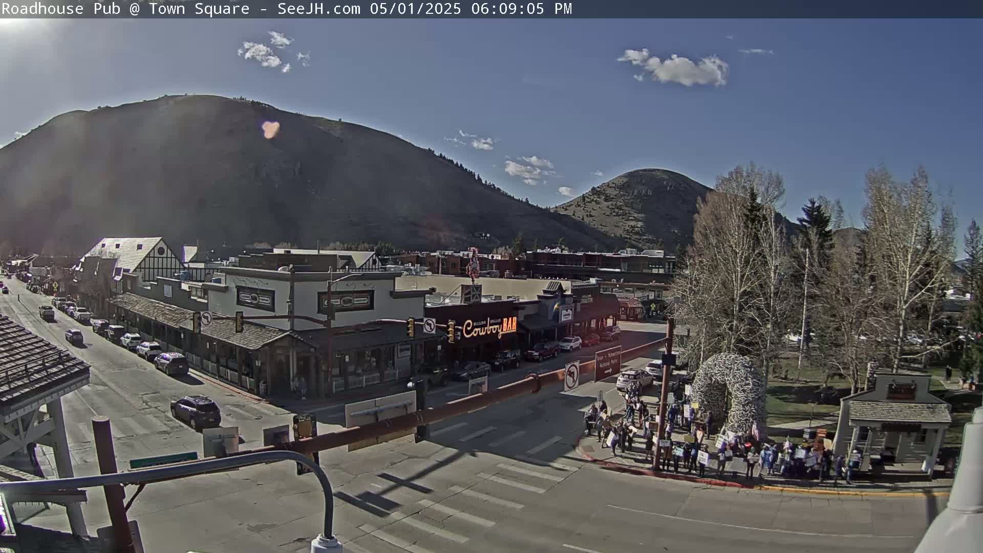 A sunny day reveals a town square with buildings, cars, and a group of people gathered near a large antler structure, all situated at the base of mountains.