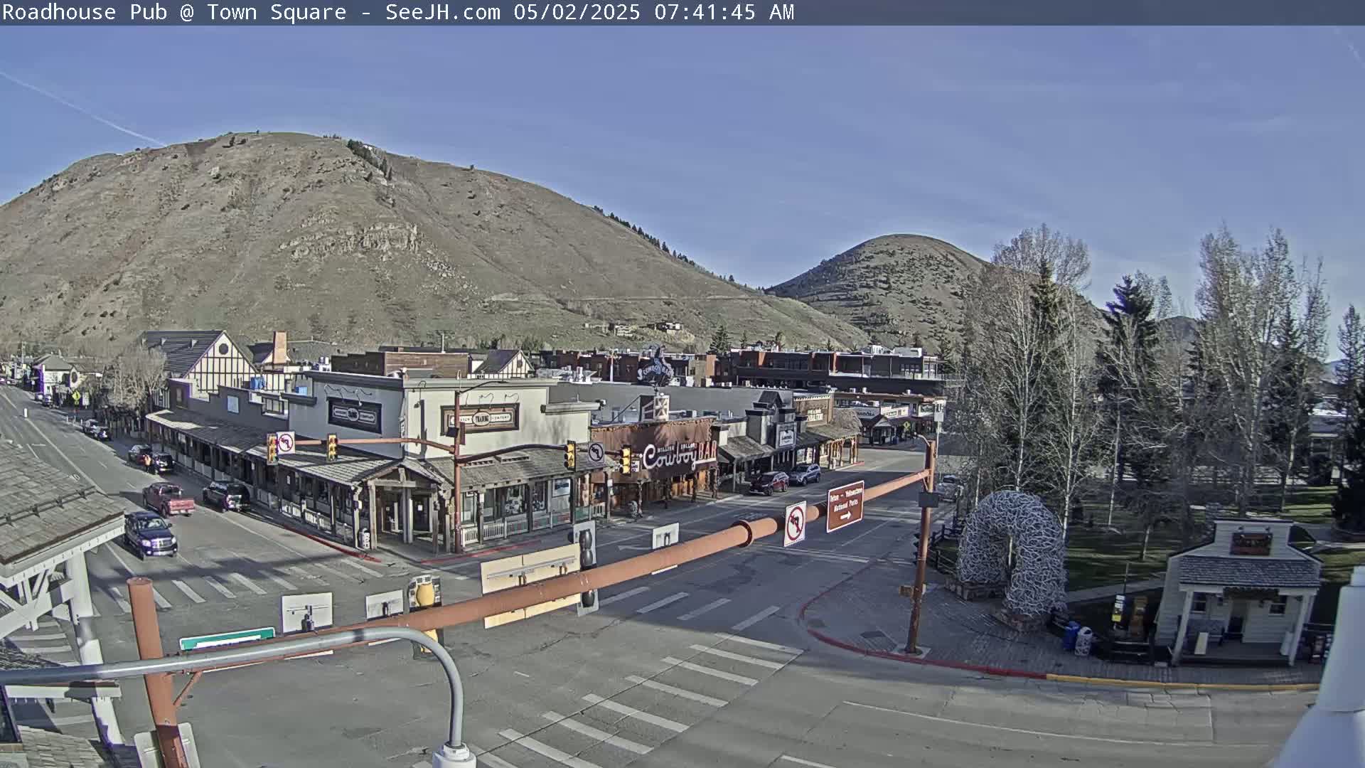 A sunny day reveals a town square with buildings, vehicles, and trees, situated at the base of two hills.