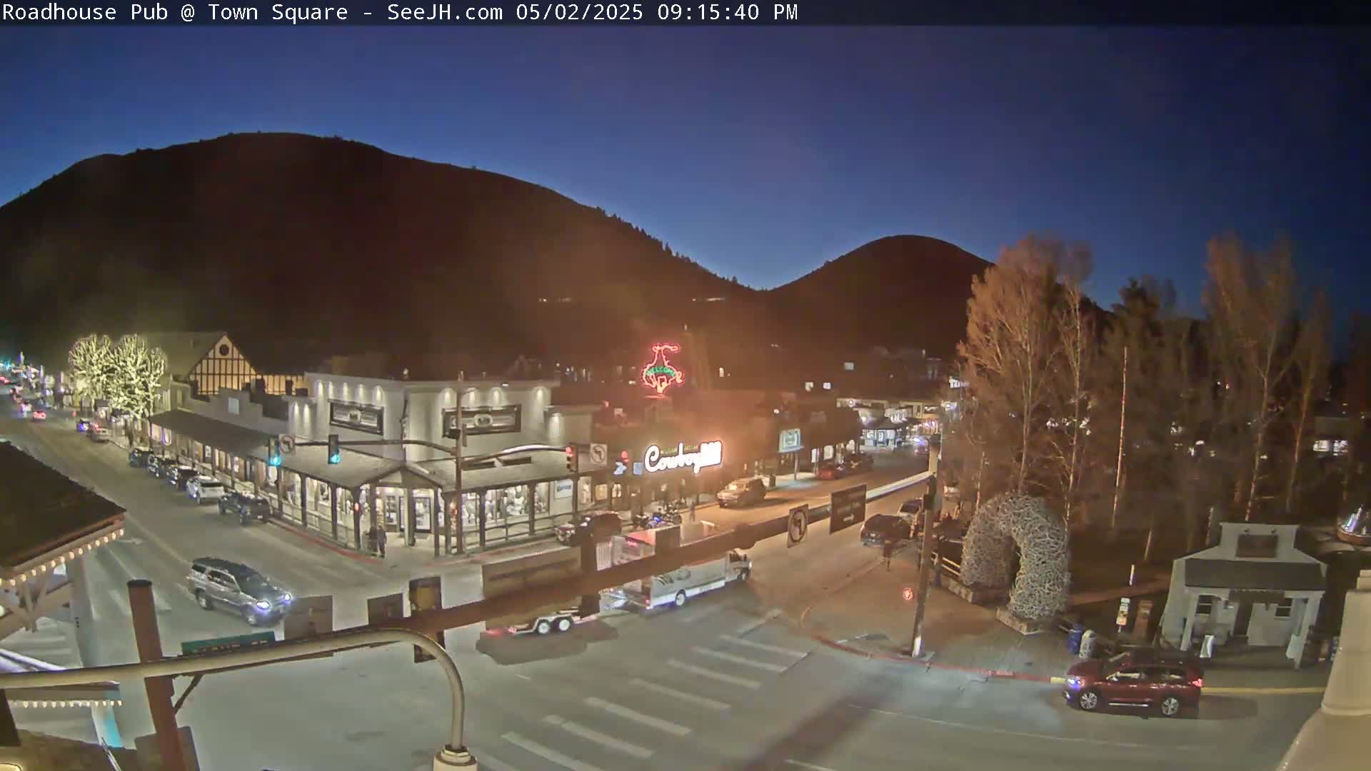 A nighttime street scene in a town nestled against dark mountains, showing sparsely trafficked streets with cars and buildings illuminated under a clear, dark blue sky.