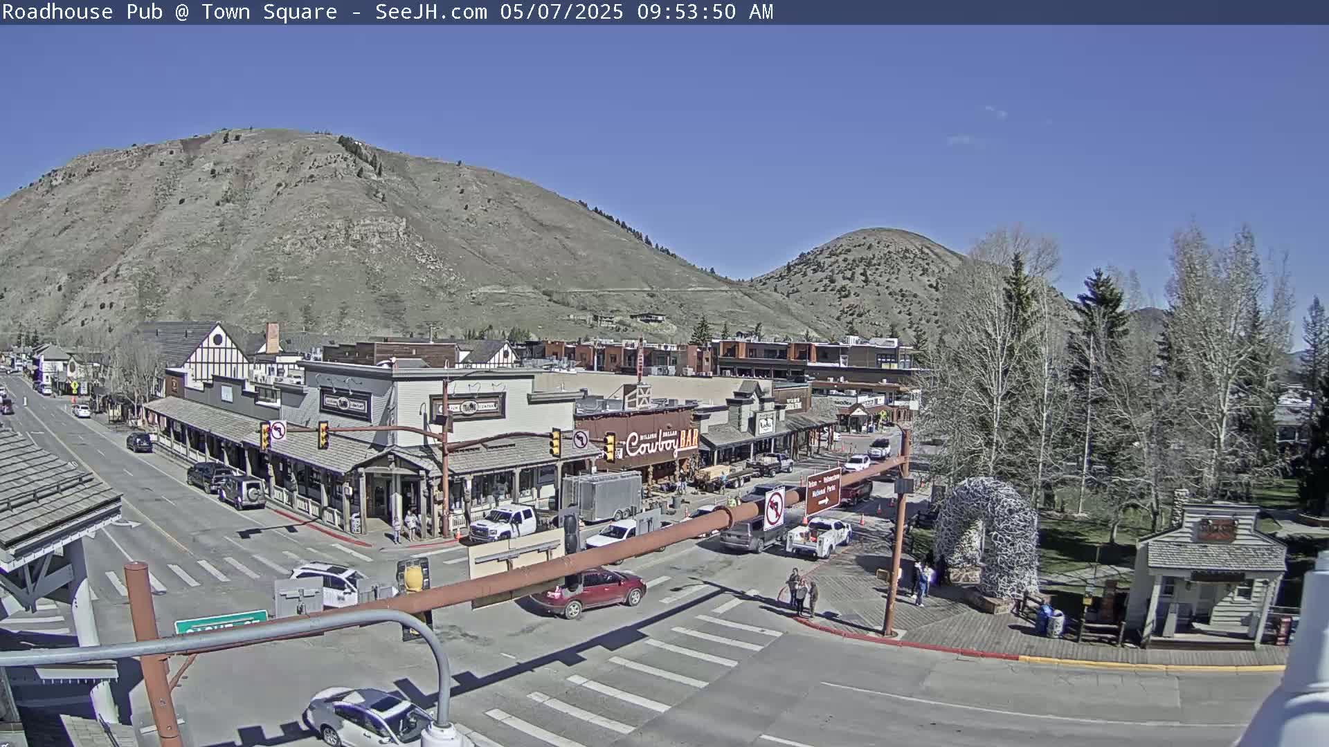 A sunny day reveals a town square nestled between mountains, with several buildings lining a street where cars are parked and driving.