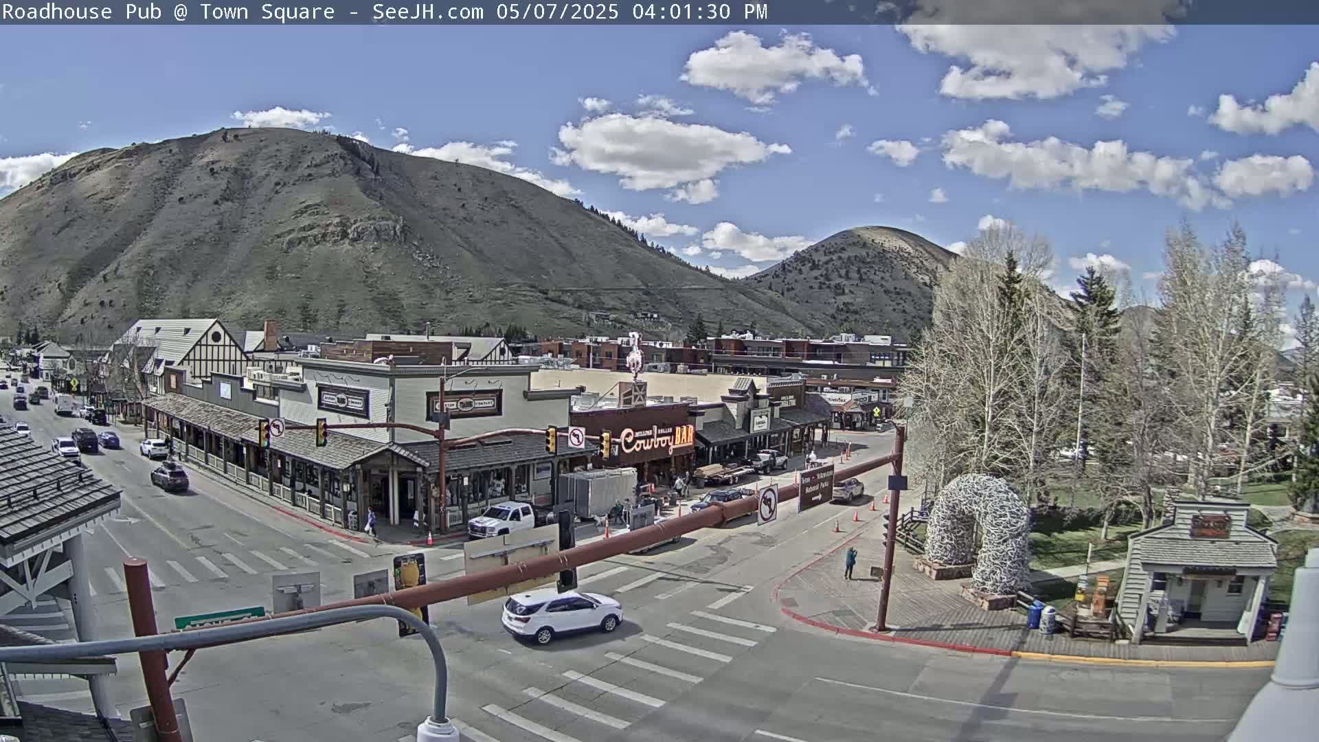 A partly cloudy day shows a mountain town square with several buildings, cars, and pedestrians, under a mostly clear blue sky.