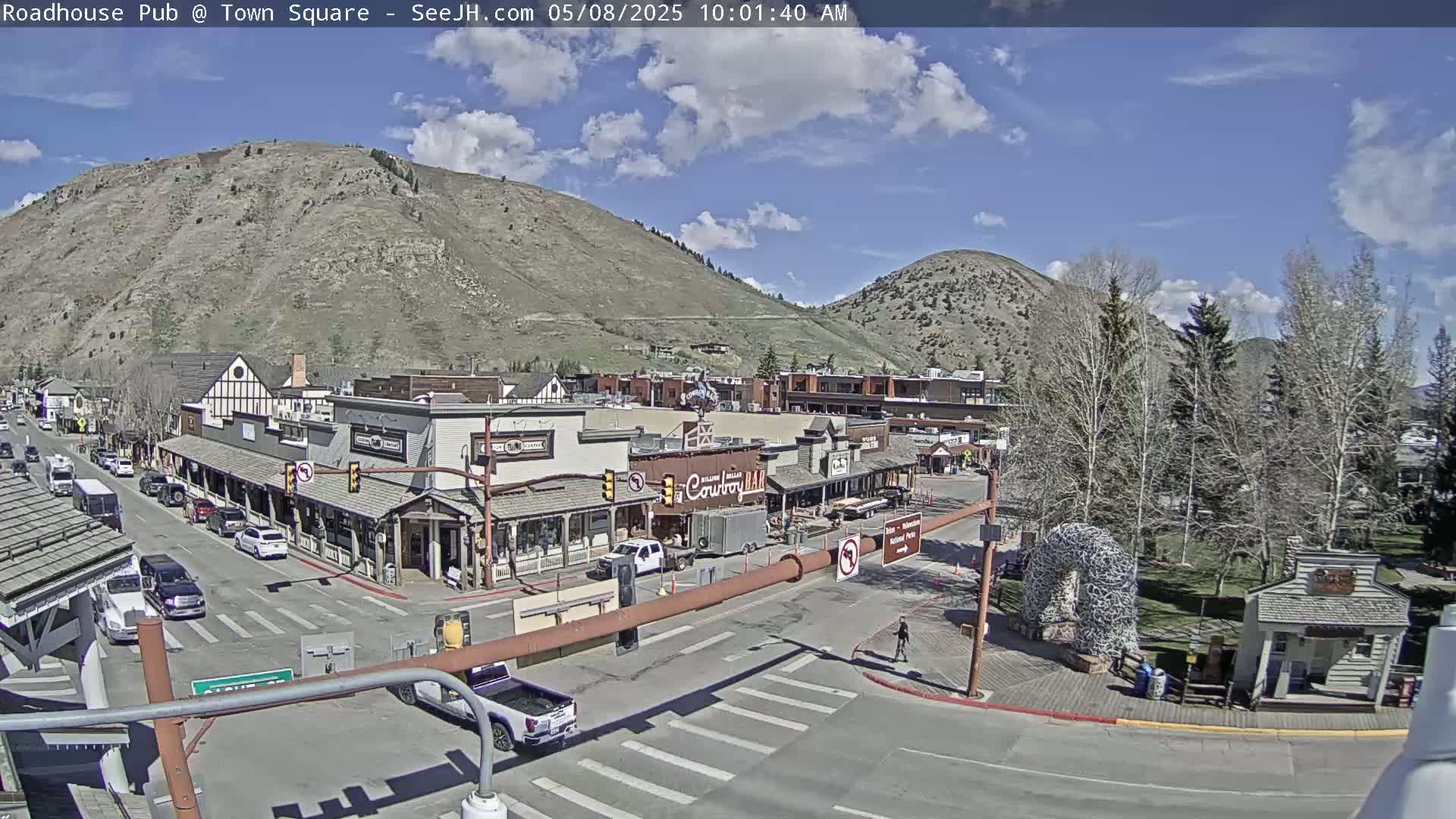 A partly sunny day shows a town square with several buildings, vehicles, and a large hill in the background.