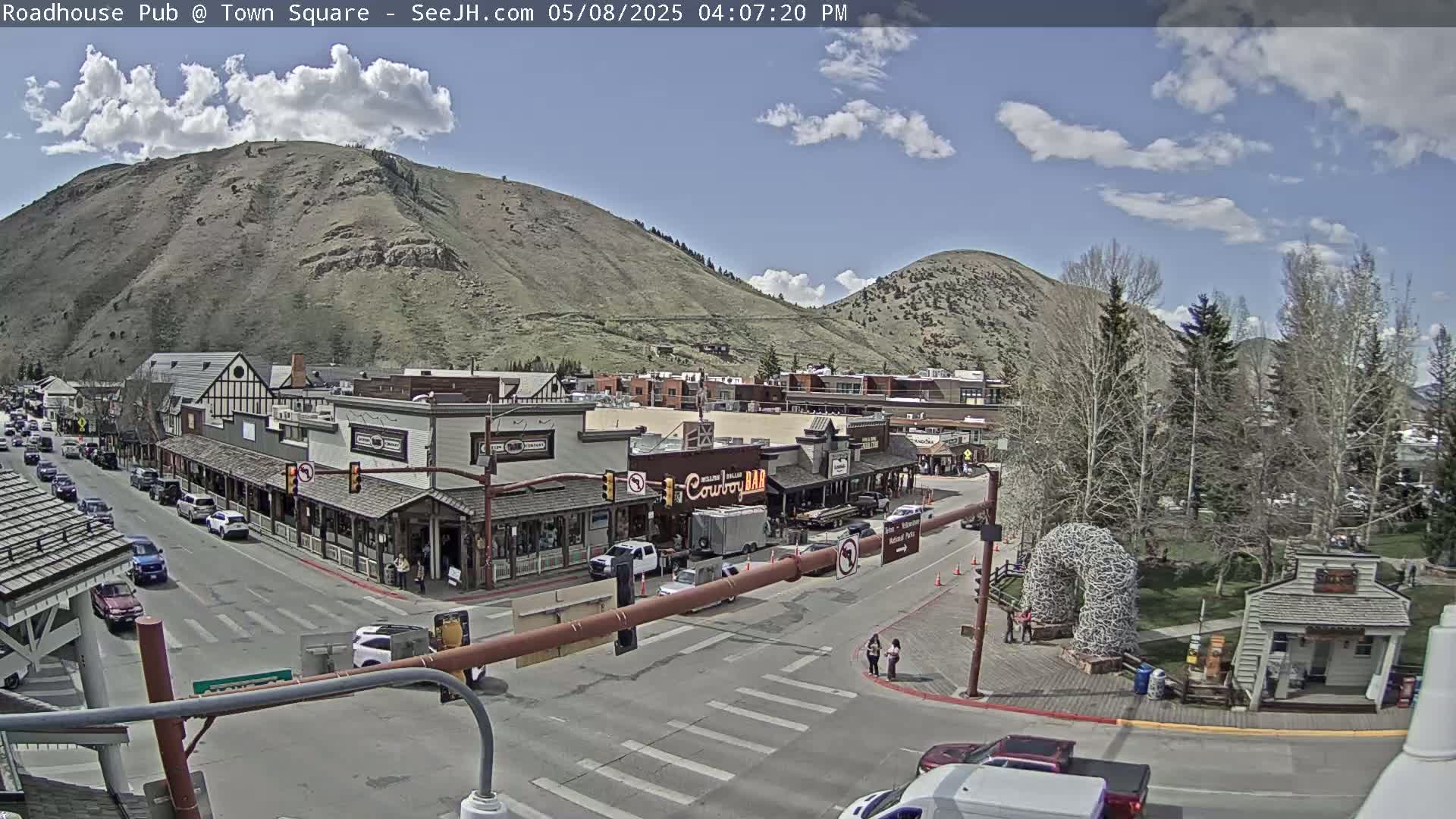 A partly cloudy day reveals a town square with shops and cars, nestled between two mountains.
