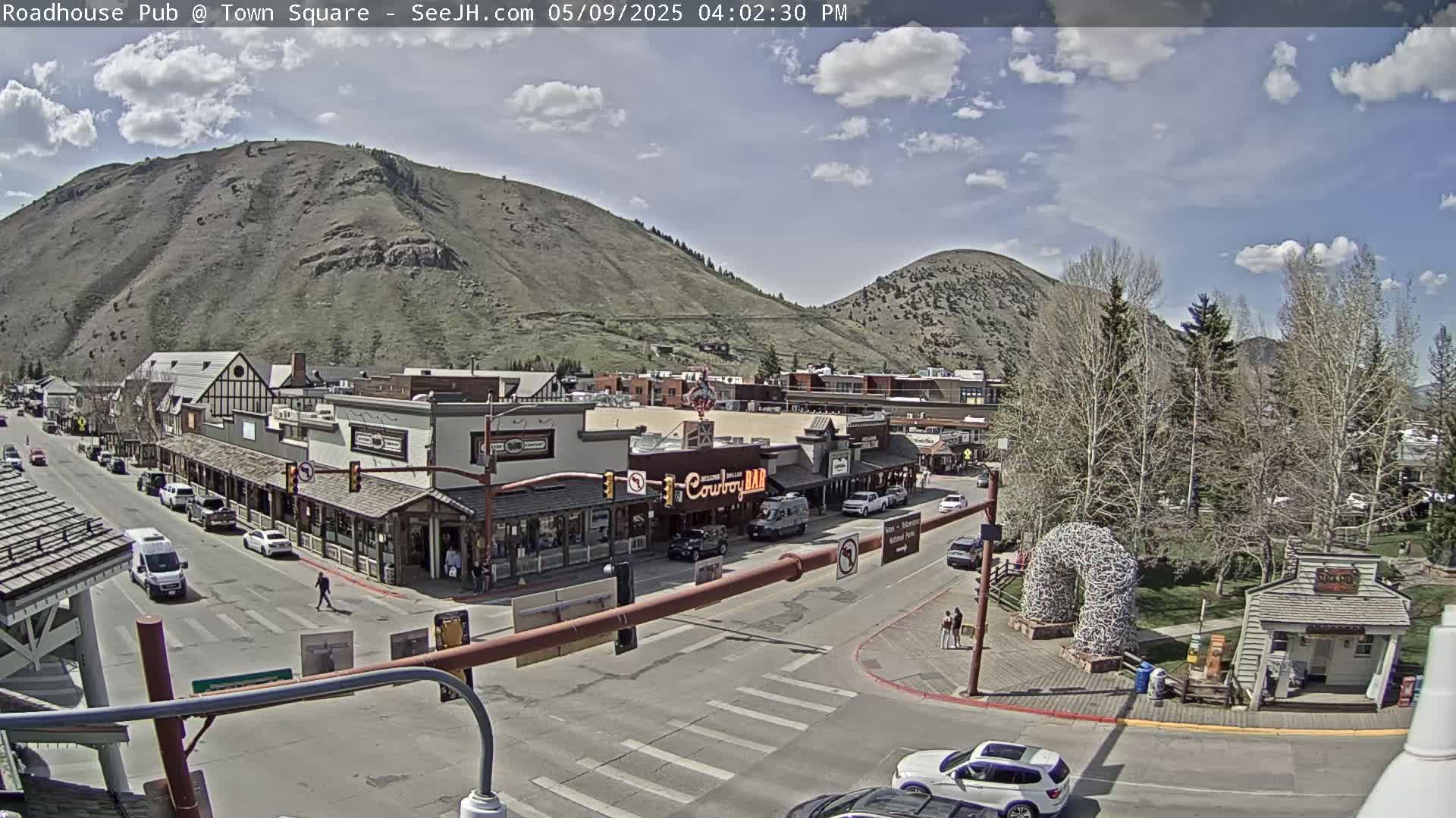 A partly sunny day reveals a town square nestled between mountains, with cars parked along the streets and pedestrians walking near shops and restaurants.