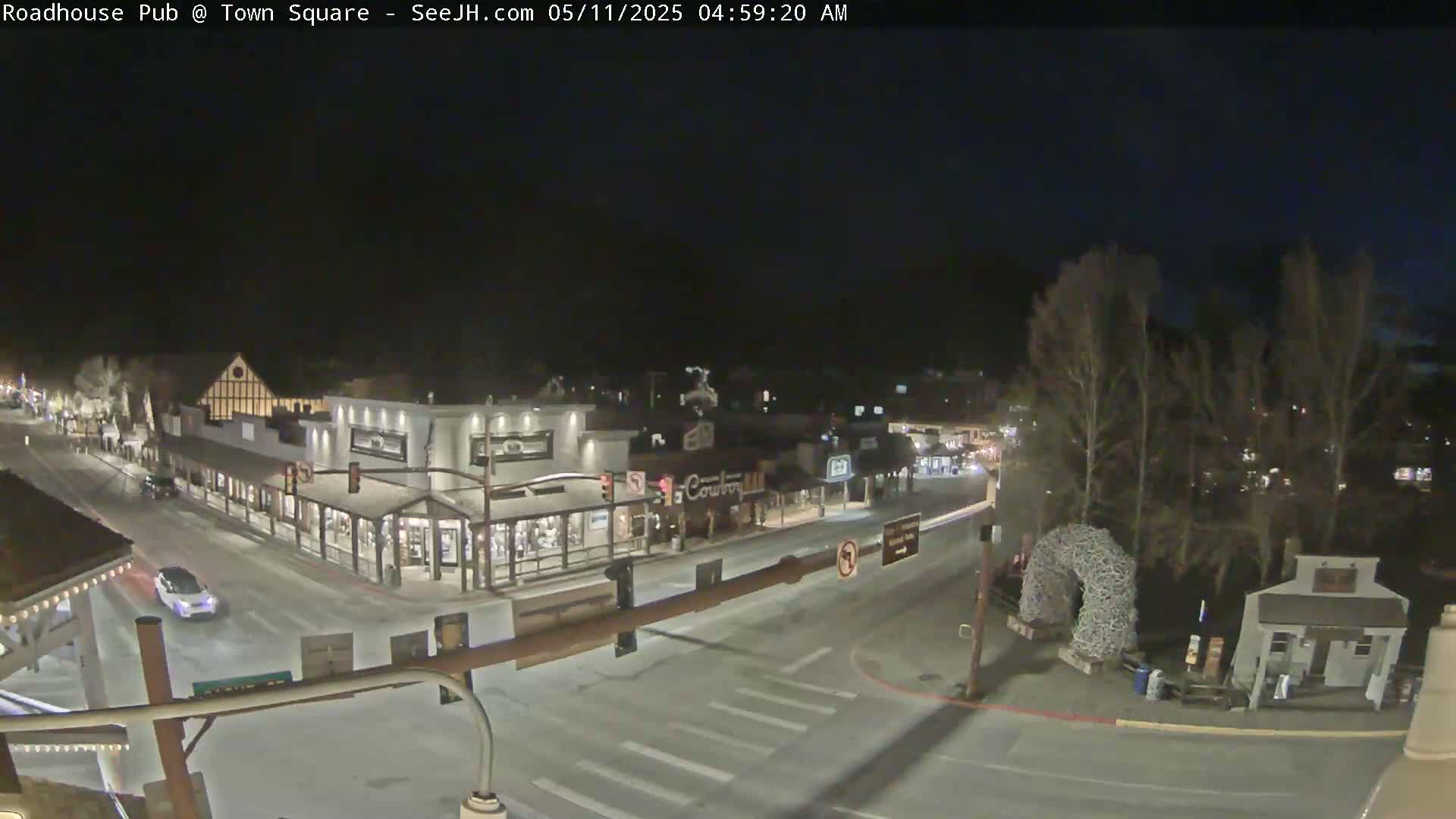 A nighttime, snow-covered town square scene shows several buildings, a few cars, and bare trees under a dark sky.