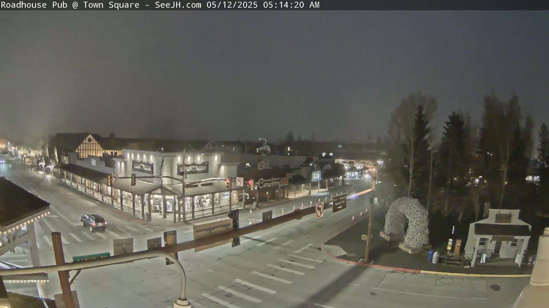 A nighttime, possibly snowy, view of a town square with a few cars and buildings visible under a dark sky.
