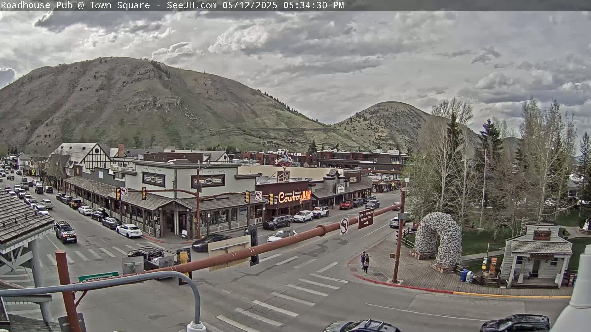 A town square with buildings, cars, and pedestrians under a cloudy sky, backed by mountains.