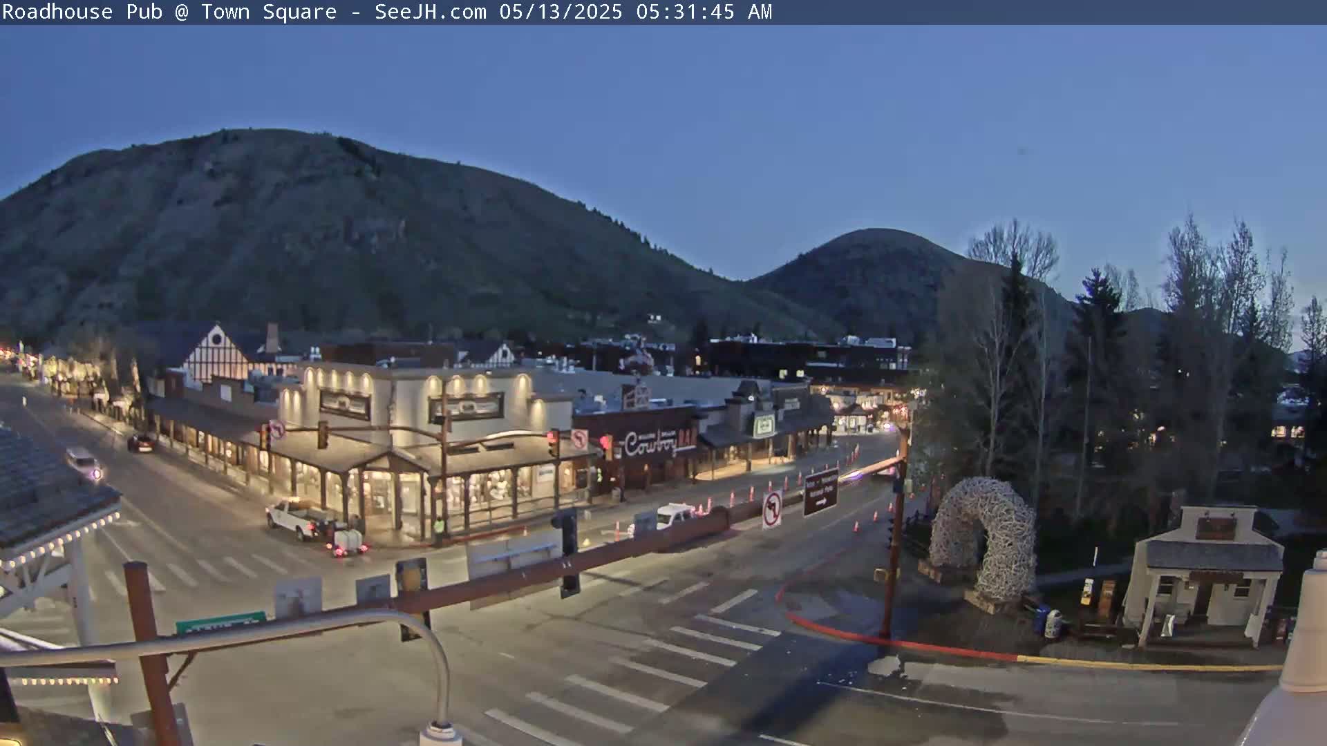 A mostly empty town square at twilight, nestled between hills, shows a few vehicles on the streets and some buildings with lights on.