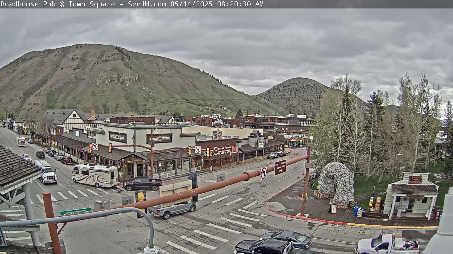 A mostly cloudy day reveals a town square with several buildings, vehicles, and a large mountain in the background.