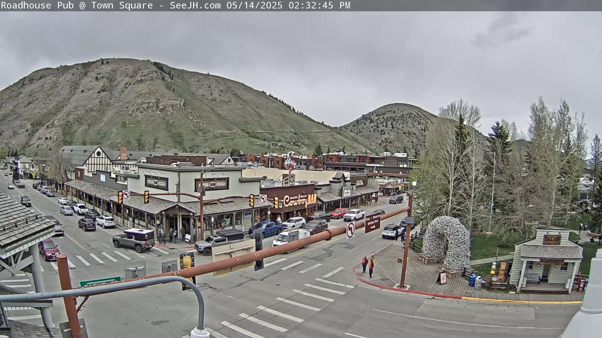 A street scene in a town nestled in a valley between two mountains on an overcast day shows buildings, cars, and pedestrians.