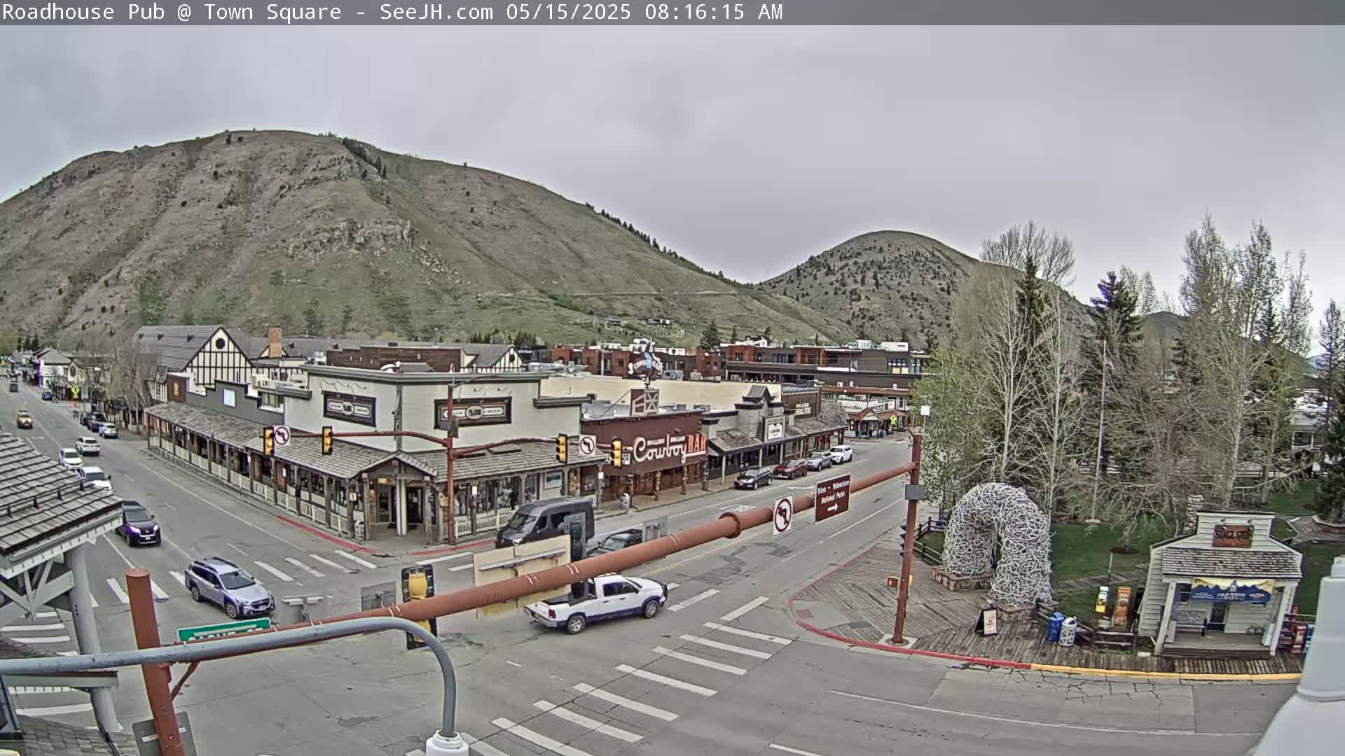 A mostly cloudy day reveals a town square nestled between mountains, with cars moving along the streets and buildings lining the sidewalks.