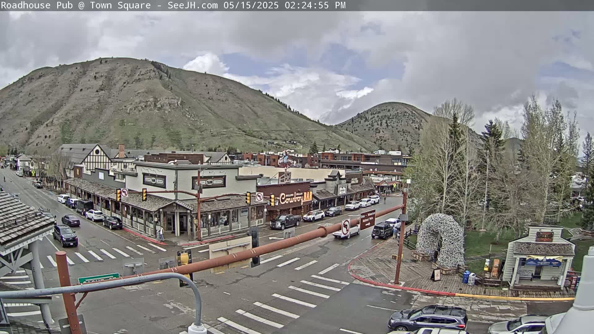 A mostly cloudy day reveals a town square with Western-style buildings, vehicles parked along the streets, and mountains in the background.