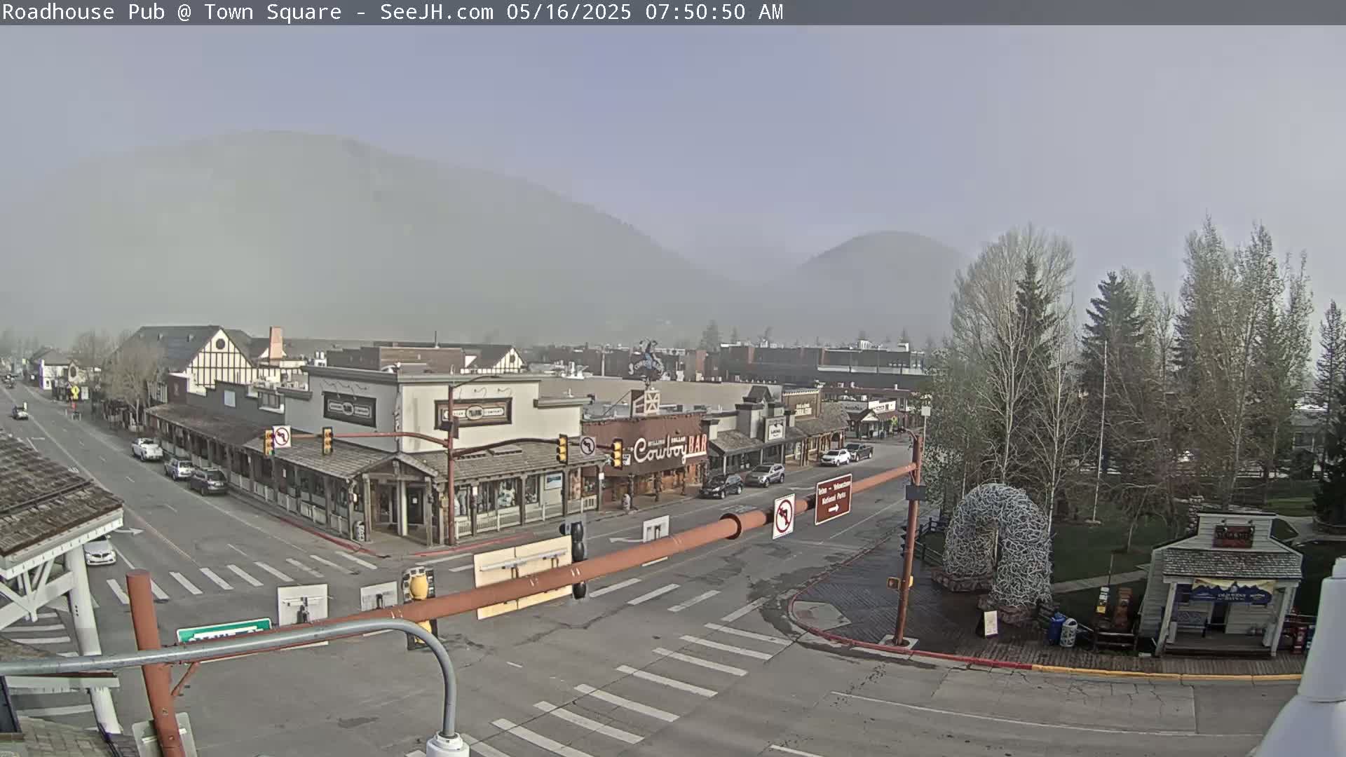 A hazy, overcast morning view of a town square with several buildings, cars on the road, and trees, all set against a backdrop of mountains.
