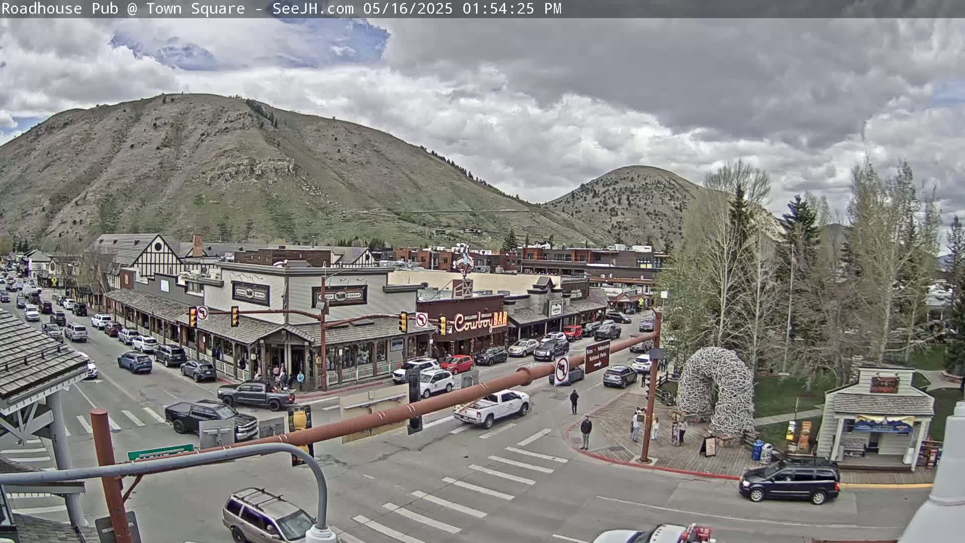 A partly cloudy day shows a town square with cars parked along the sides of the street,  buildings lining the street, and a large hill behind it.