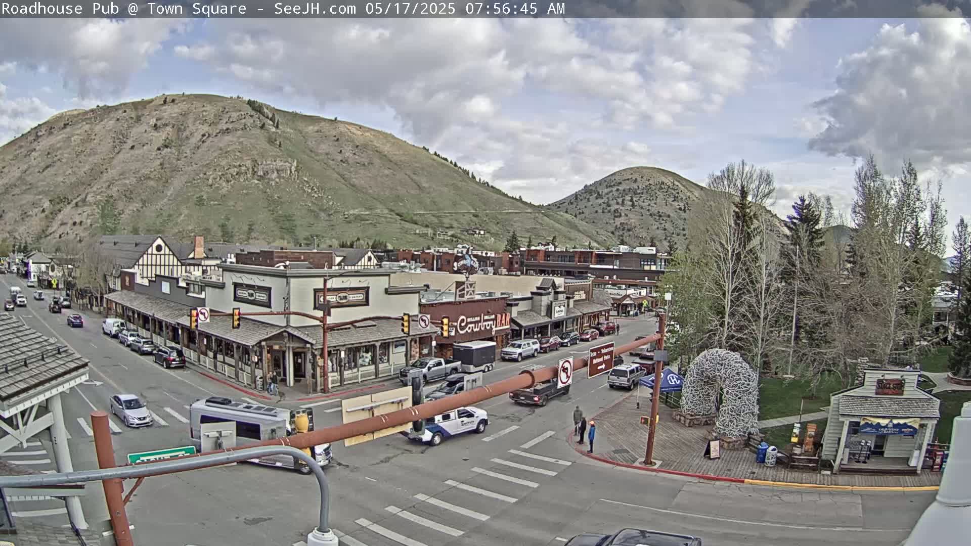 A partly cloudy morning scene shows a town square with several buildings, cars parked along the street, and a few people walking around, all nestled against a backdrop of mountains.