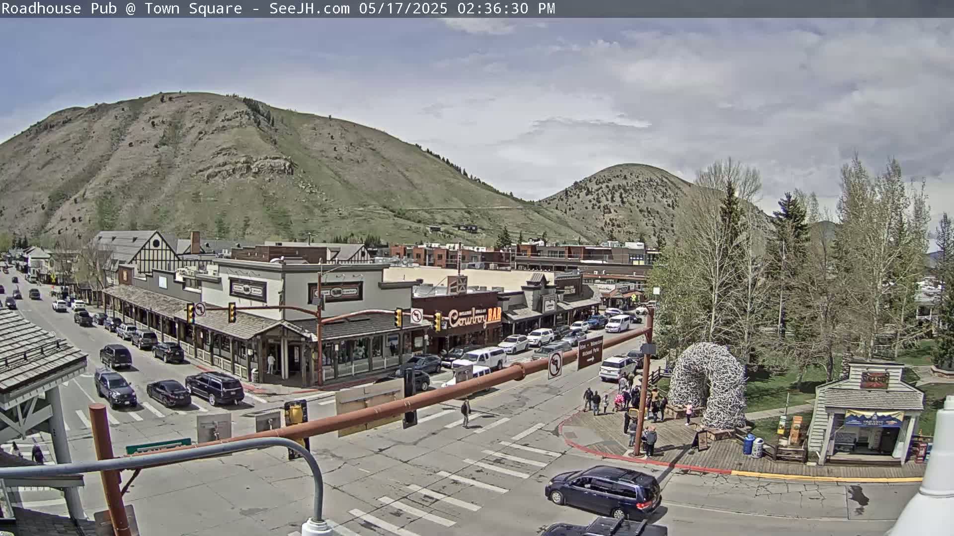 A partly cloudy afternoon scene shows a town square with buildings, cars, and pedestrians, nestled at the base of a mountain.