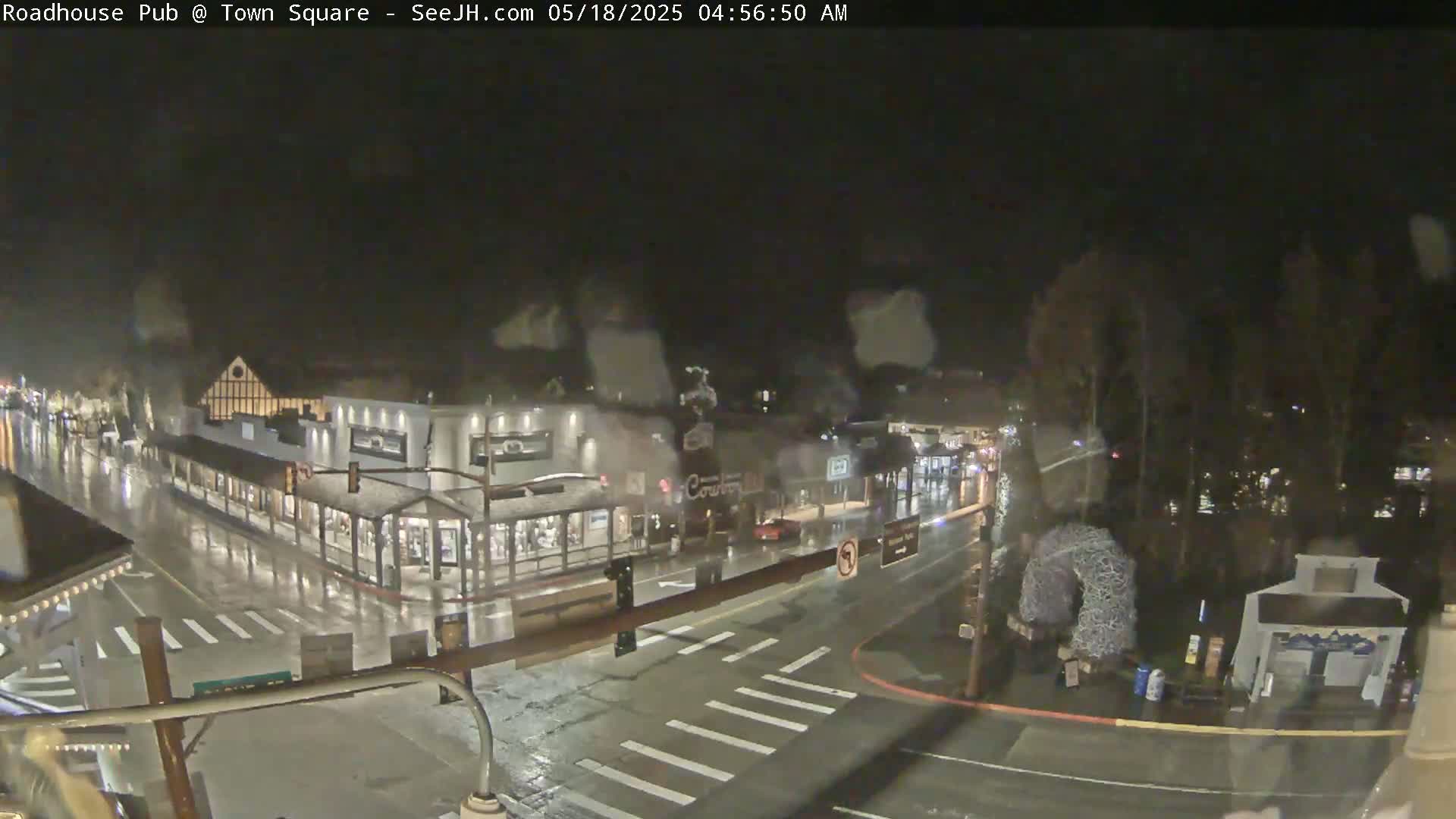 A rain-slicked town square at night, showing a street intersection and several buildings under dim streetlights.