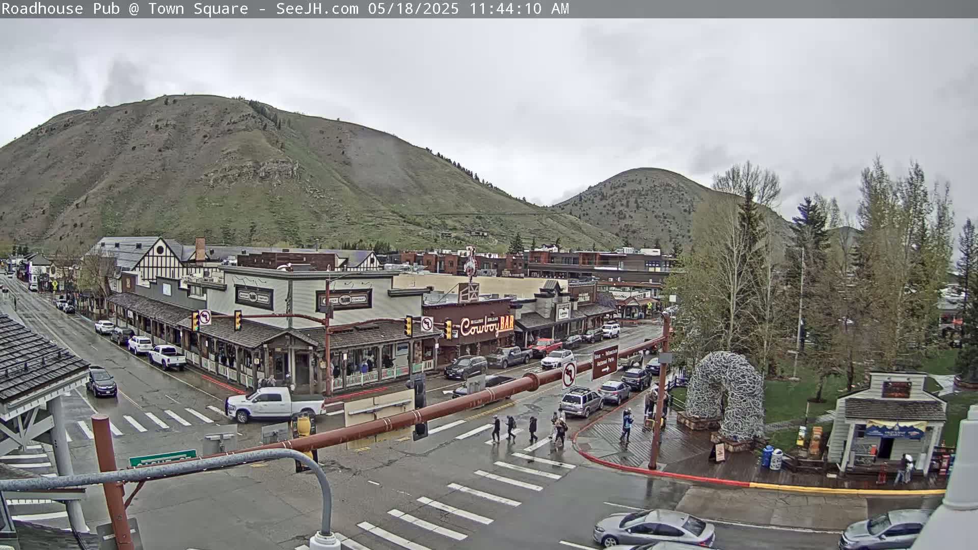 A wet town square with pedestrians crossing a street, surrounded by buildings nestled at the base of a mountain under an overcast sky.