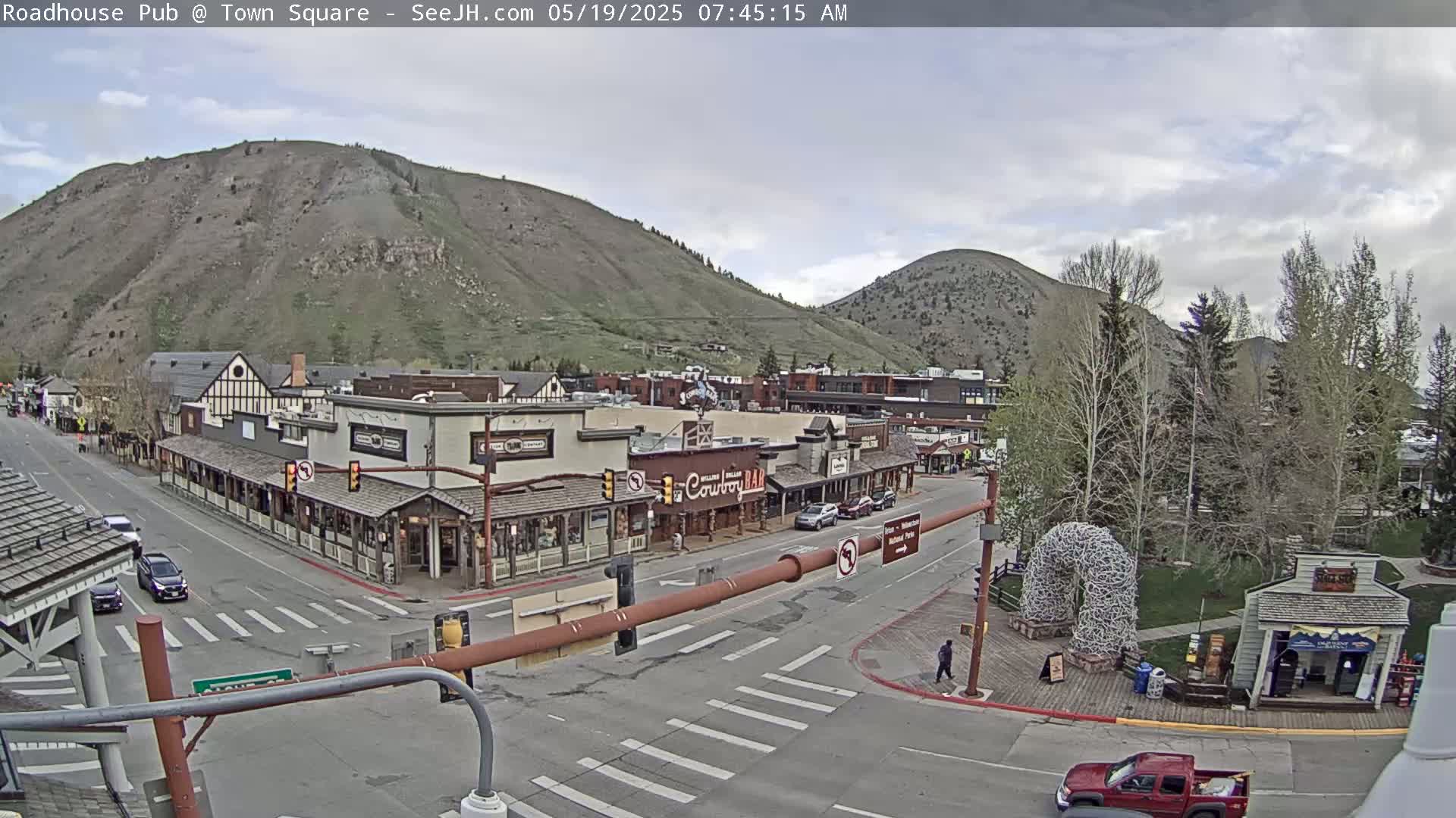 A mostly cloudy day reveals a mountain town square with several buildings, cars, and pedestrians, alongside a large antler arch.