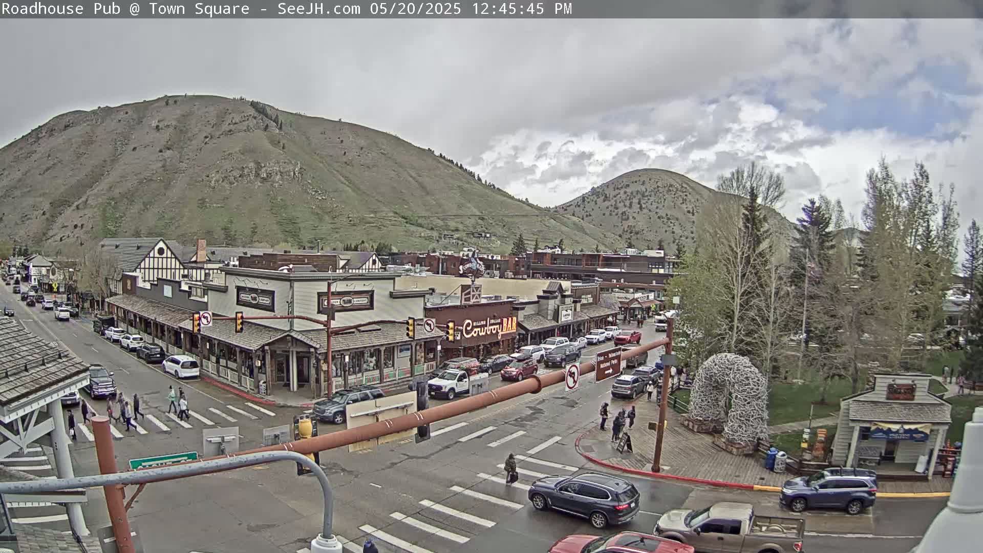 A partly cloudy day reveals a mountain town square with pedestrians crossing a street, cars parked along the sides of buildings, and several shops lining the street.