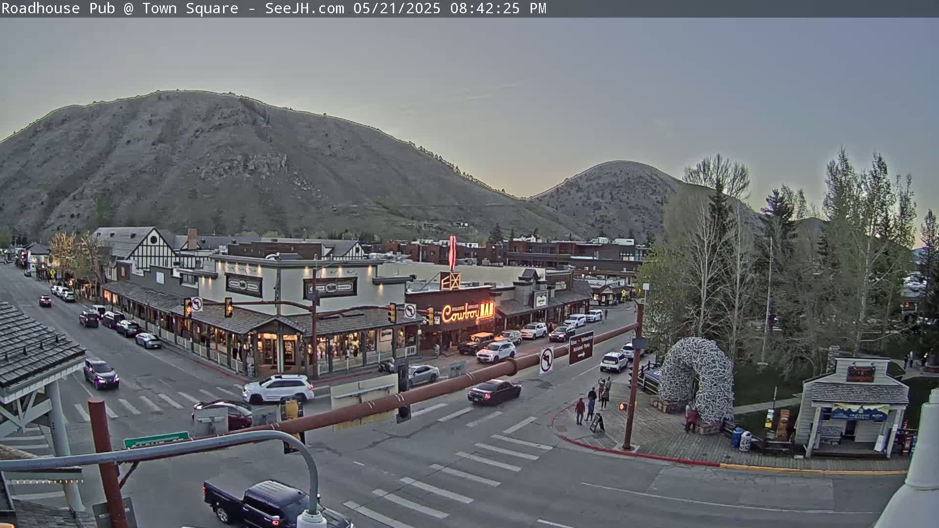 A twilight view of a town square with several buildings, cars, and people, situated at the base of a large hill under a clear sky.