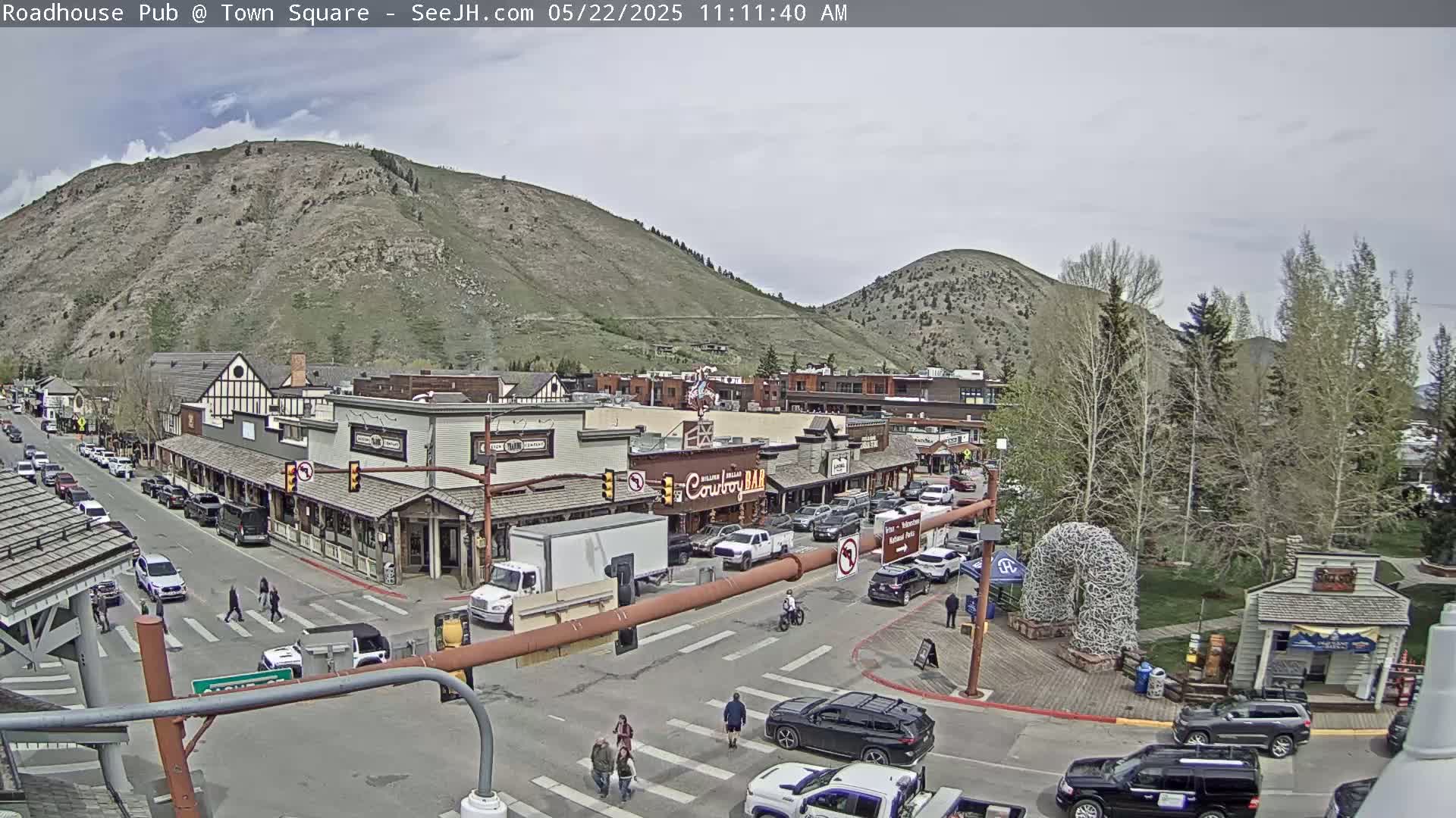 A partly cloudy day in a town square surrounded by mountains, featuring several buildings, cars, and pedestrians crossing a street.