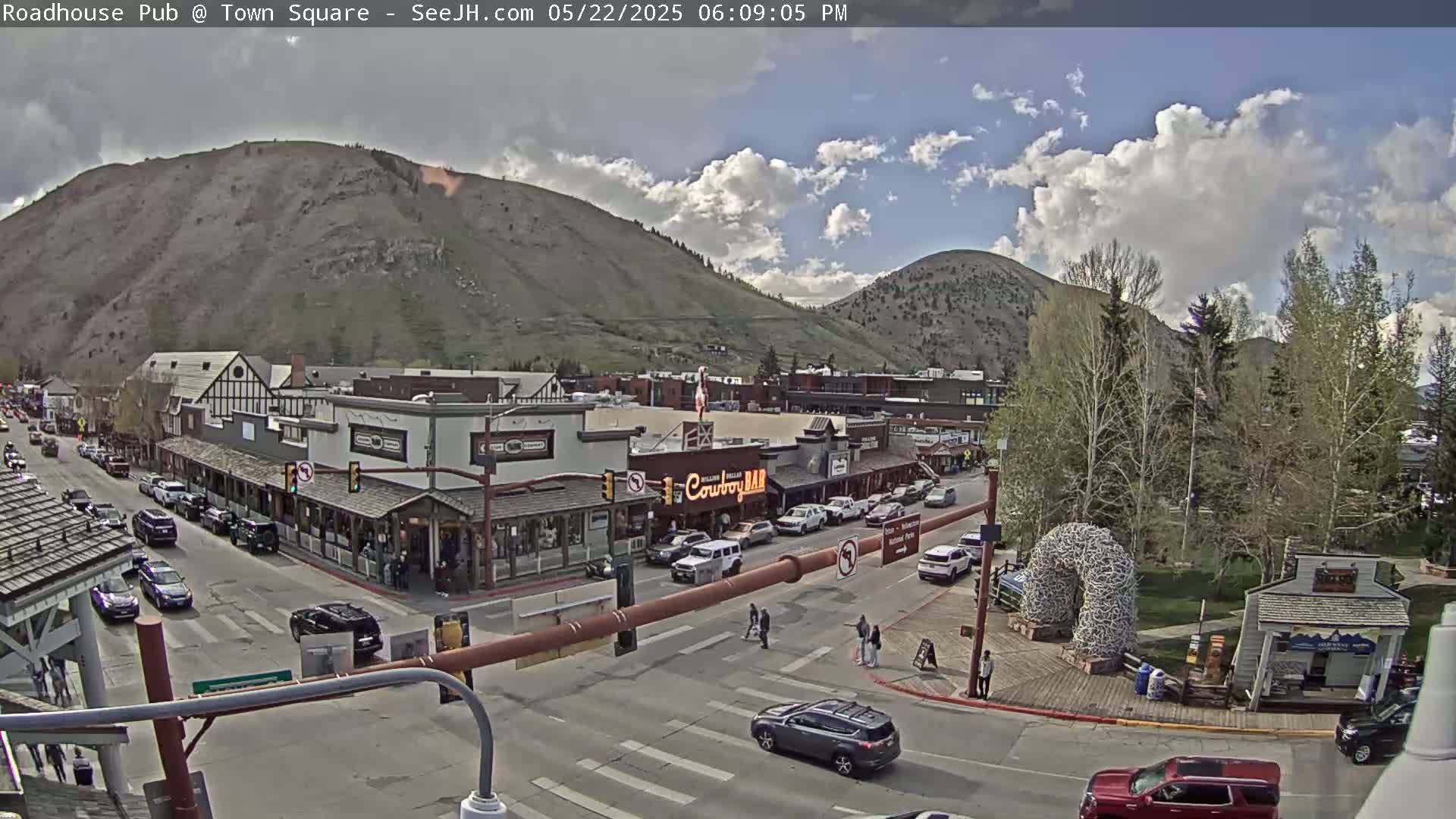 A partly cloudy afternoon scene shows a mountain town square with cars parked and driving on streets, buildings lining the streets, and people walking on the sidewalks, all nestled at the foot of a mountain.