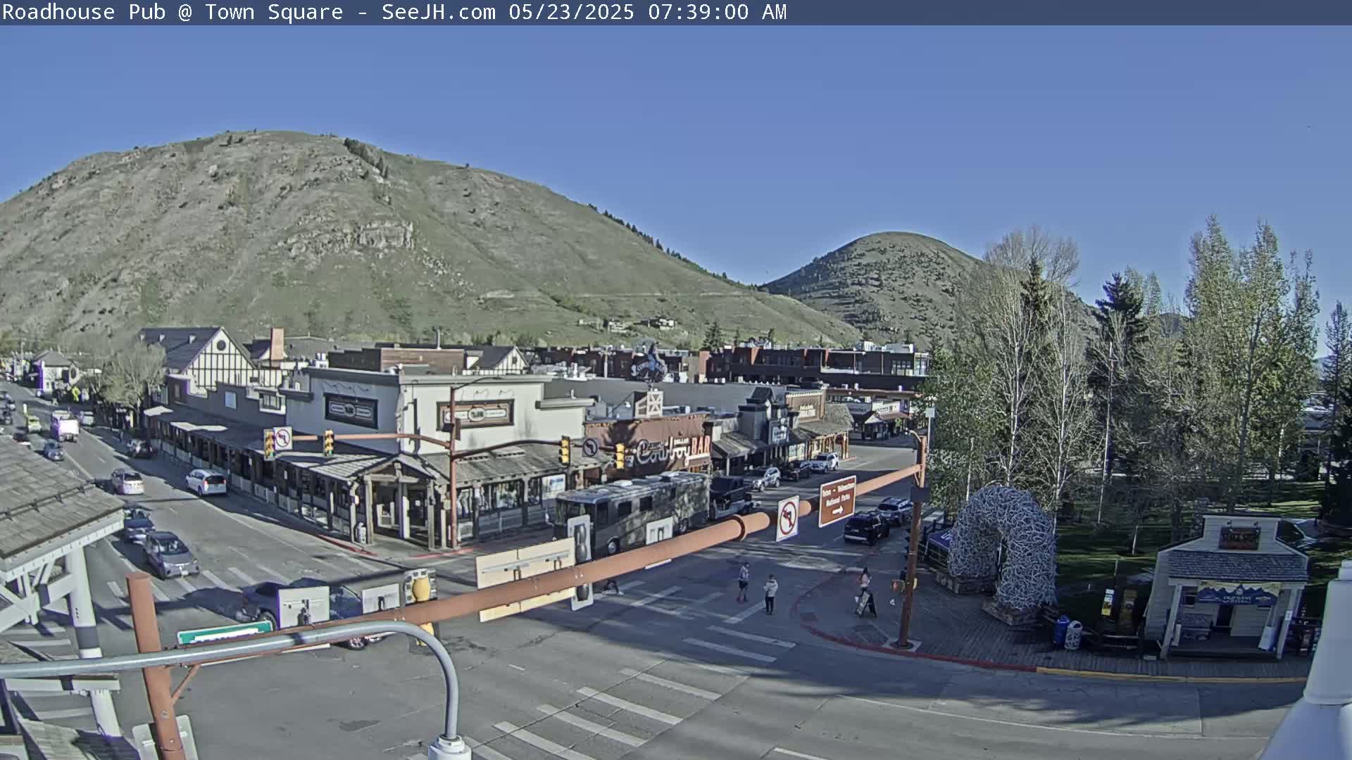 A street scene in a town nestled between hills on a sunny day shows vehicles on roads intersecting near buildings and pedestrians crossing the street.
