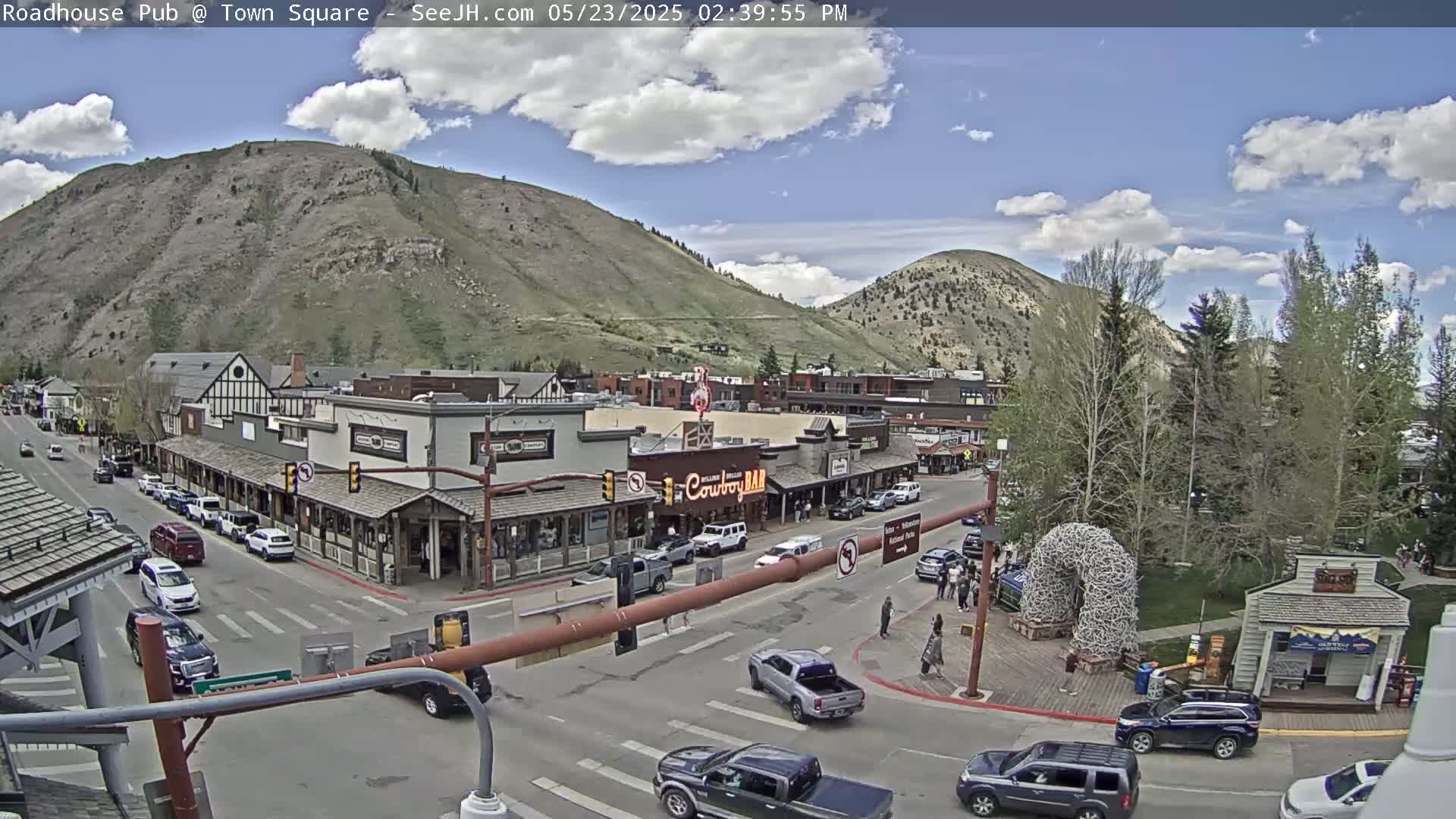 A partly cloudy day reveals a mountain town square with cars on the streets, buildings lining the sides, and trees in a nearby park.