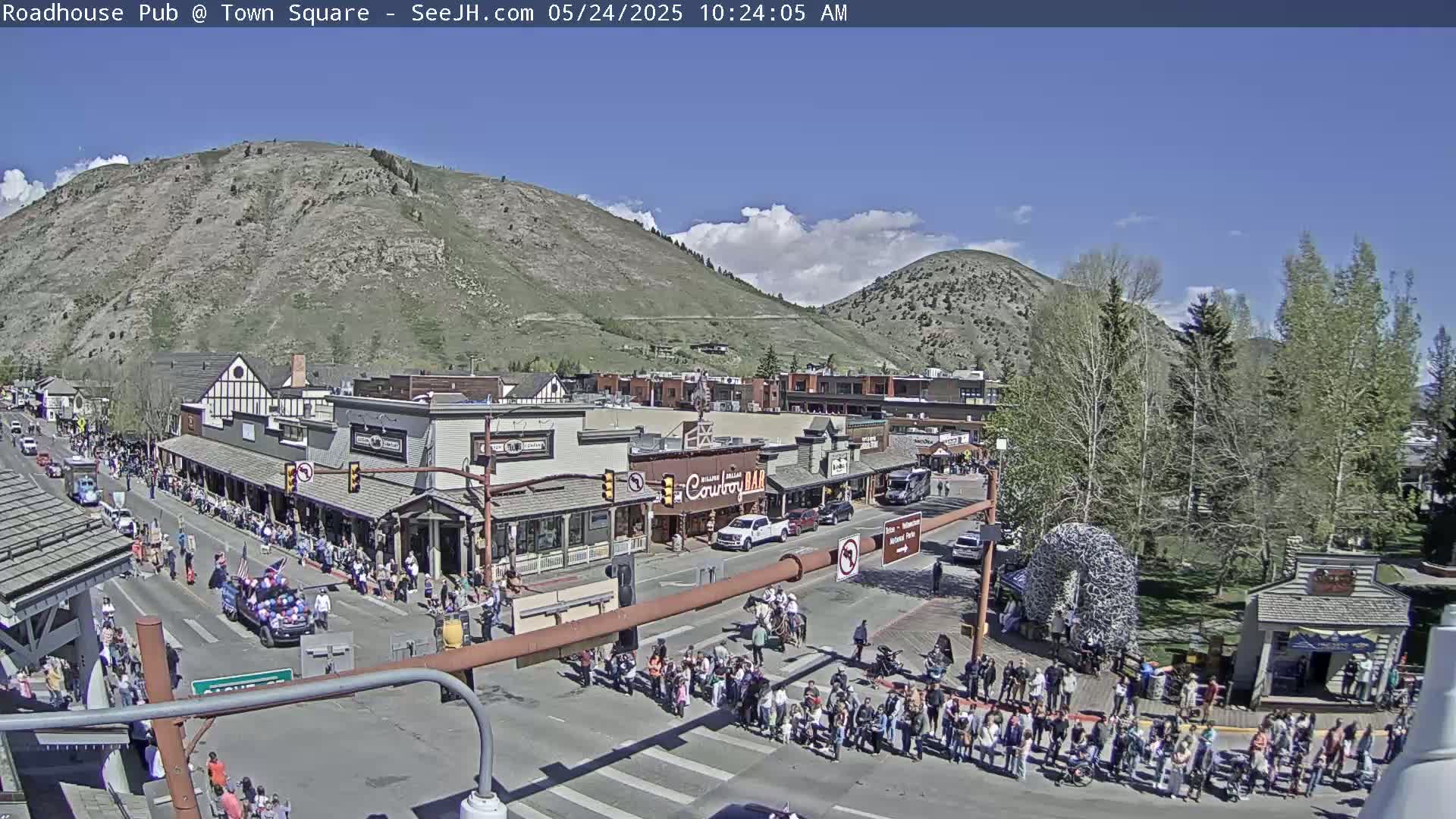 A sunny day with some clouds shows a crowd of people lining a town square street, with a parade passing by in front of buildings nestled against a mountainside.