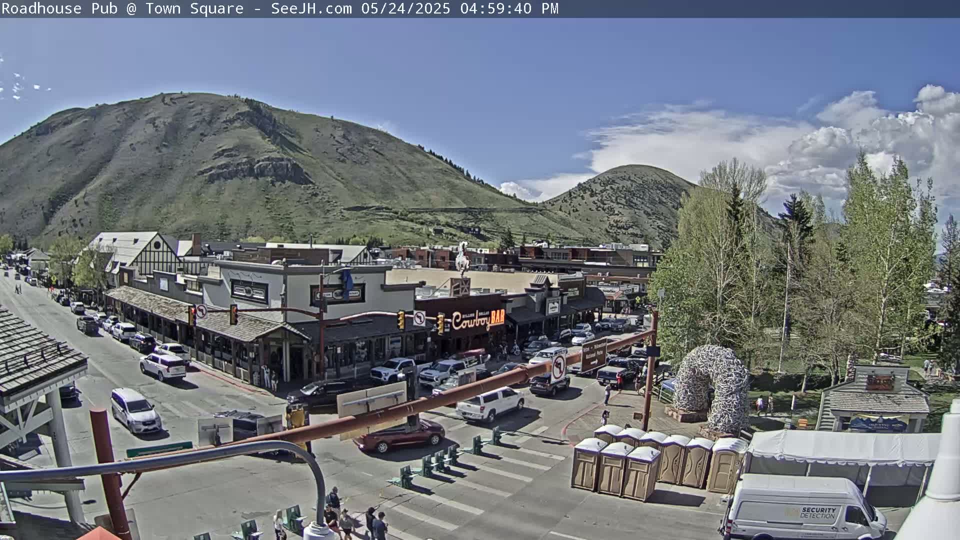 A sunny day shows a town square with cars parked along the street, buildings lining the sides, and a mountain in the background.