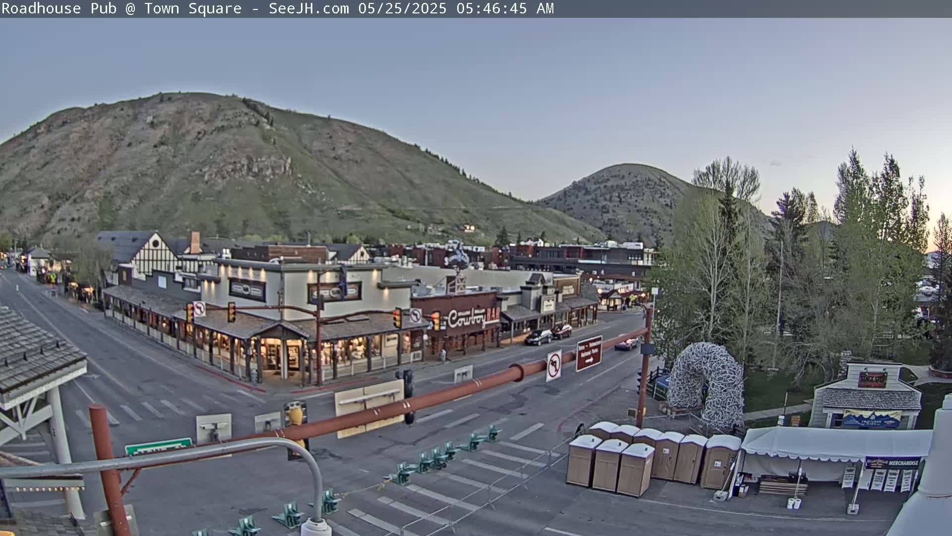 A mostly clear dawn illuminates a town square nestled between two hills, with several buildings lining a street and a few vehicles visible.