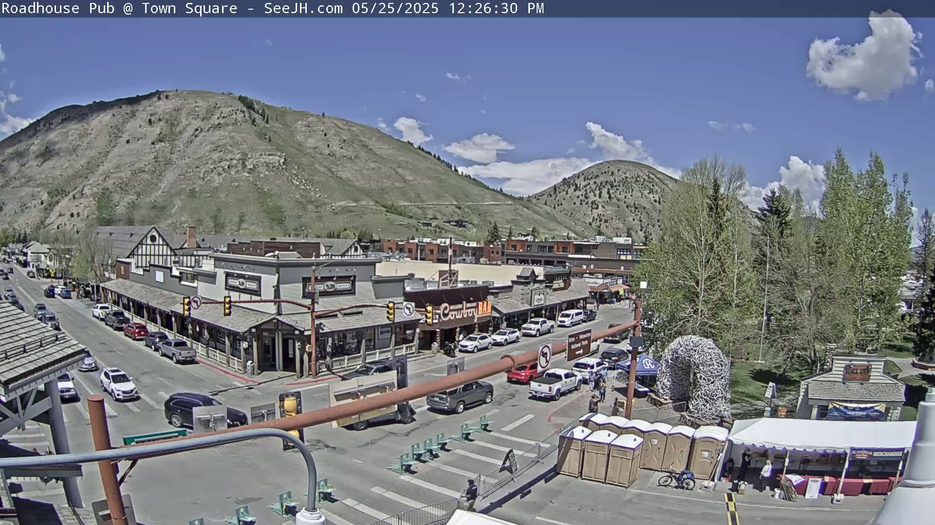 A partly sunny day in a mountain town shows a street lined with shops and buildings, with cars parked along the side and a few people walking around.