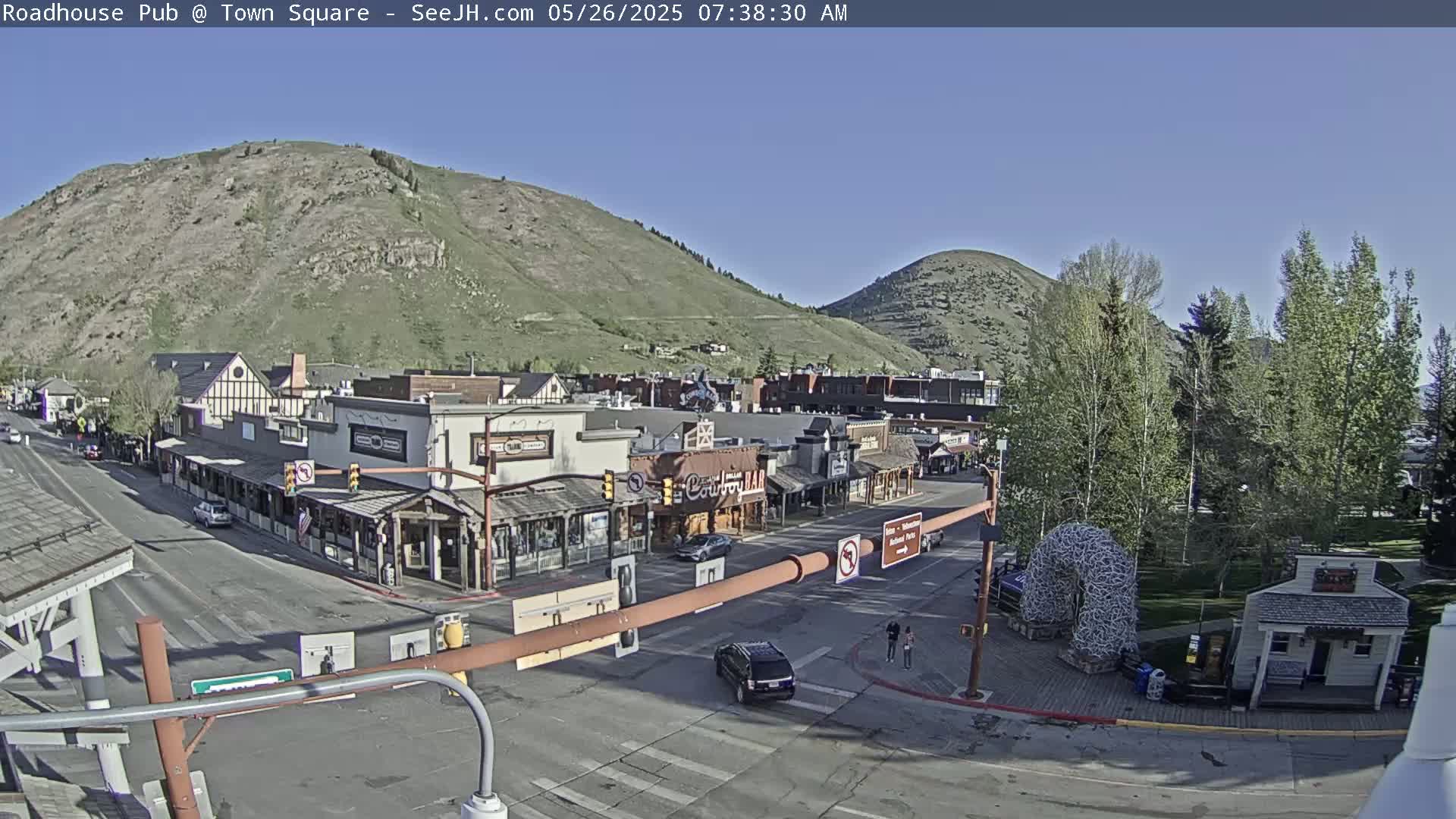 A sunny morning view of a town square nestled between mountains, with several buildings lining a street intersection, and a few vehicles and pedestrians visible.