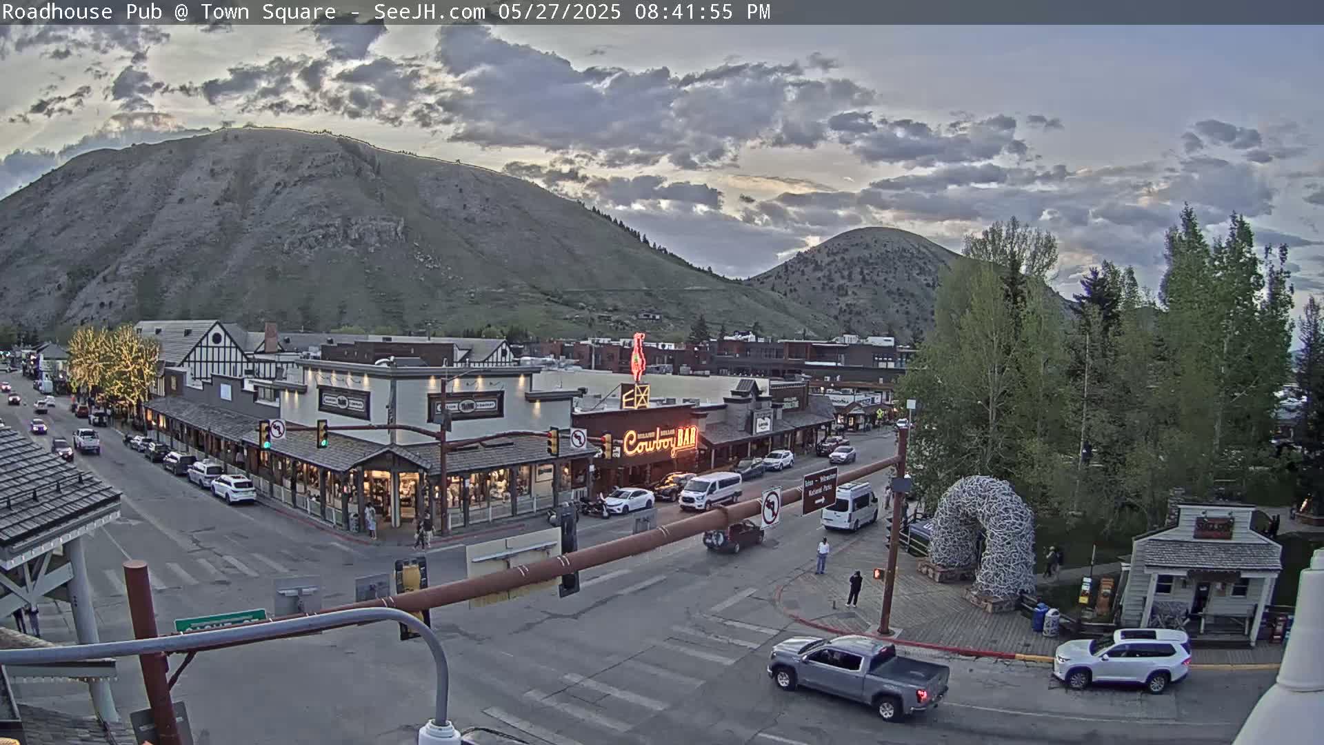 A mountain town street scene at dusk under partly cloudy skies shows cars parked and driving on a street lined with shops and buildings, a large hill in the background.