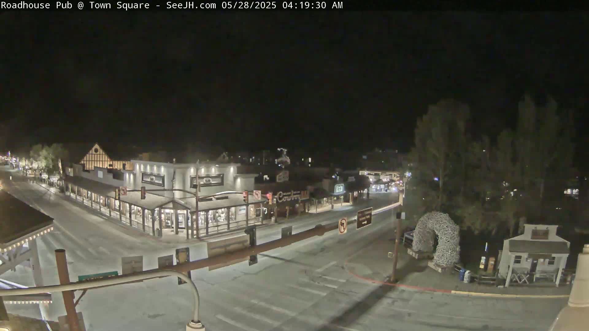 A nighttime, snow-covered town square with several buildings, streetlights, and a large antler archway is visible from an elevated vantage point.