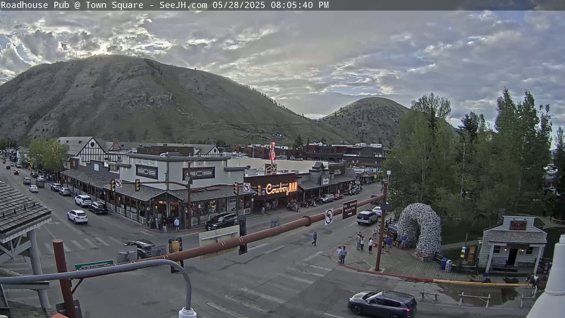 A partly cloudy late afternoon scene shows a town square with buildings, cars, and pedestrians, nestled against a backdrop of mountains.