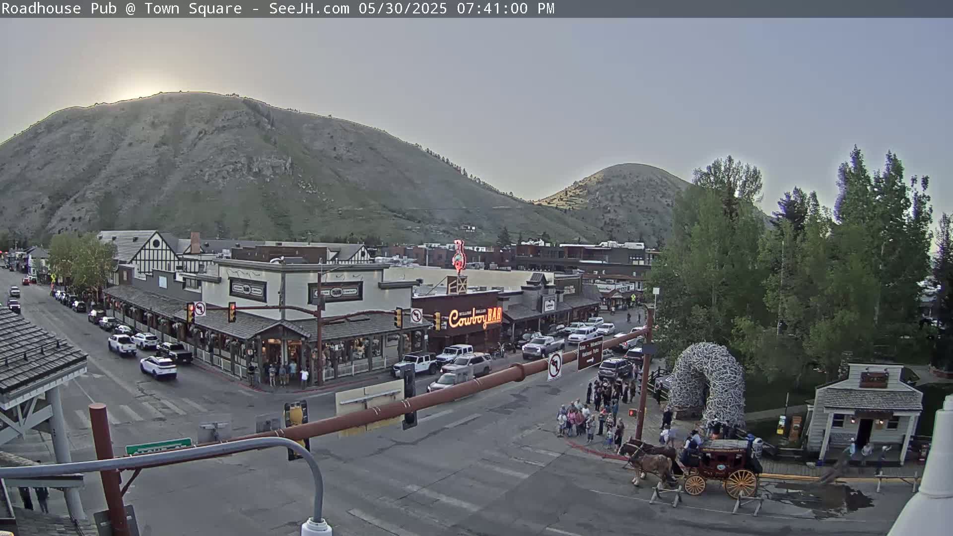 A high-angle, evening view of a town square nestled between mountains, featuring buildings, vehicles, pedestrians, and a horse-drawn carriage under a clear sky.
