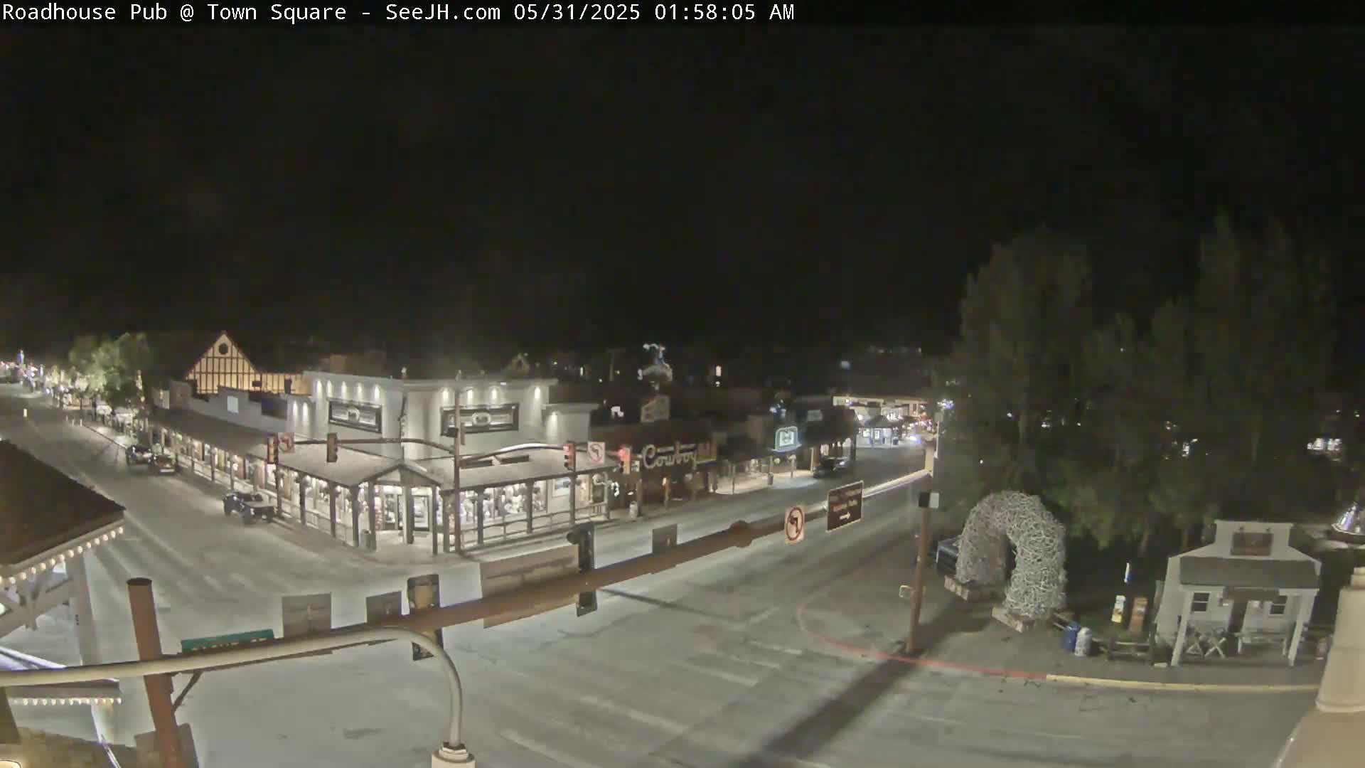 A nighttime view of a snow-covered town square with several buildings and vehicles, illuminated by streetlights.