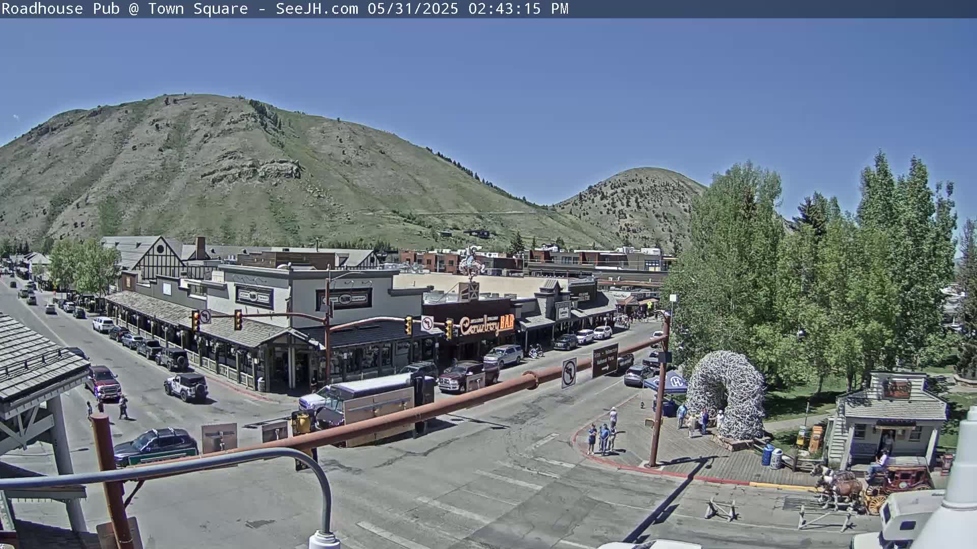 A sunny town square scene shows buildings, vehicles, and people, situated at the base of a mountain range under a clear blue sky.